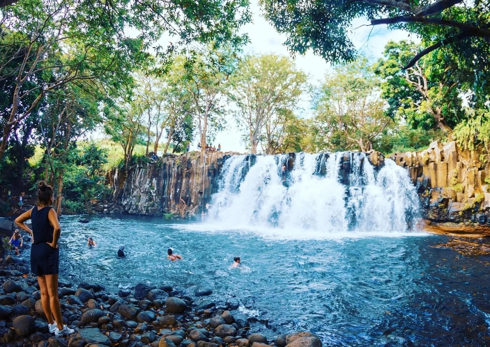 People swimming and standing by a waterfall in a lush, green forest.