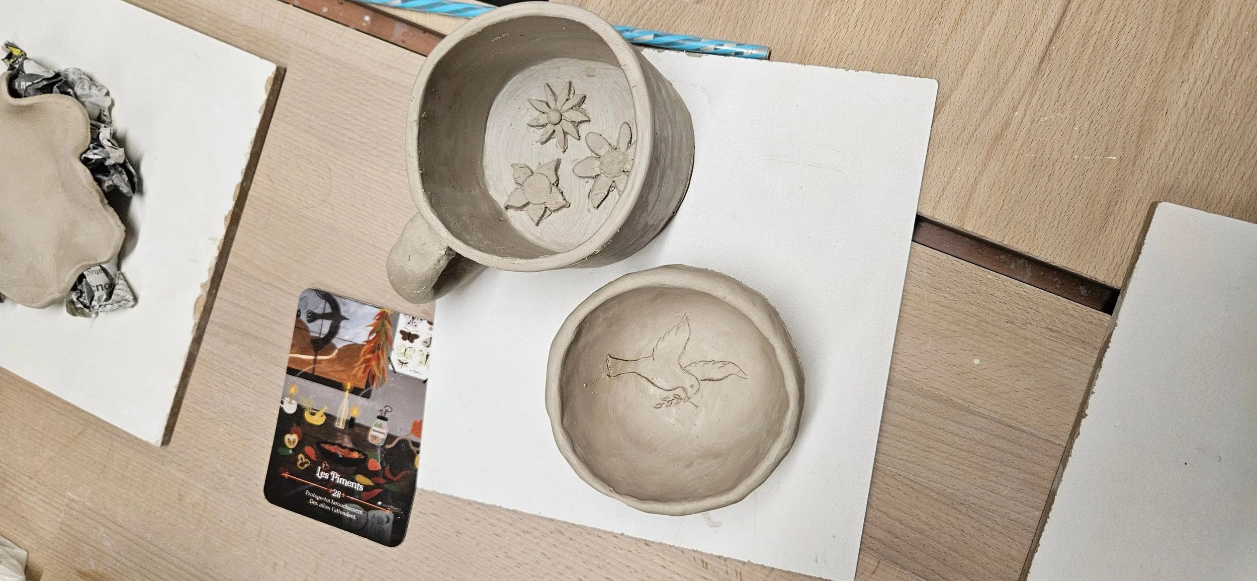 Deux céramiques en cours de création, une tasse avec des motifs floraux et une petite assiette avec un oiseau, posées sur des supports en papier et sur un plan de travail en bois.