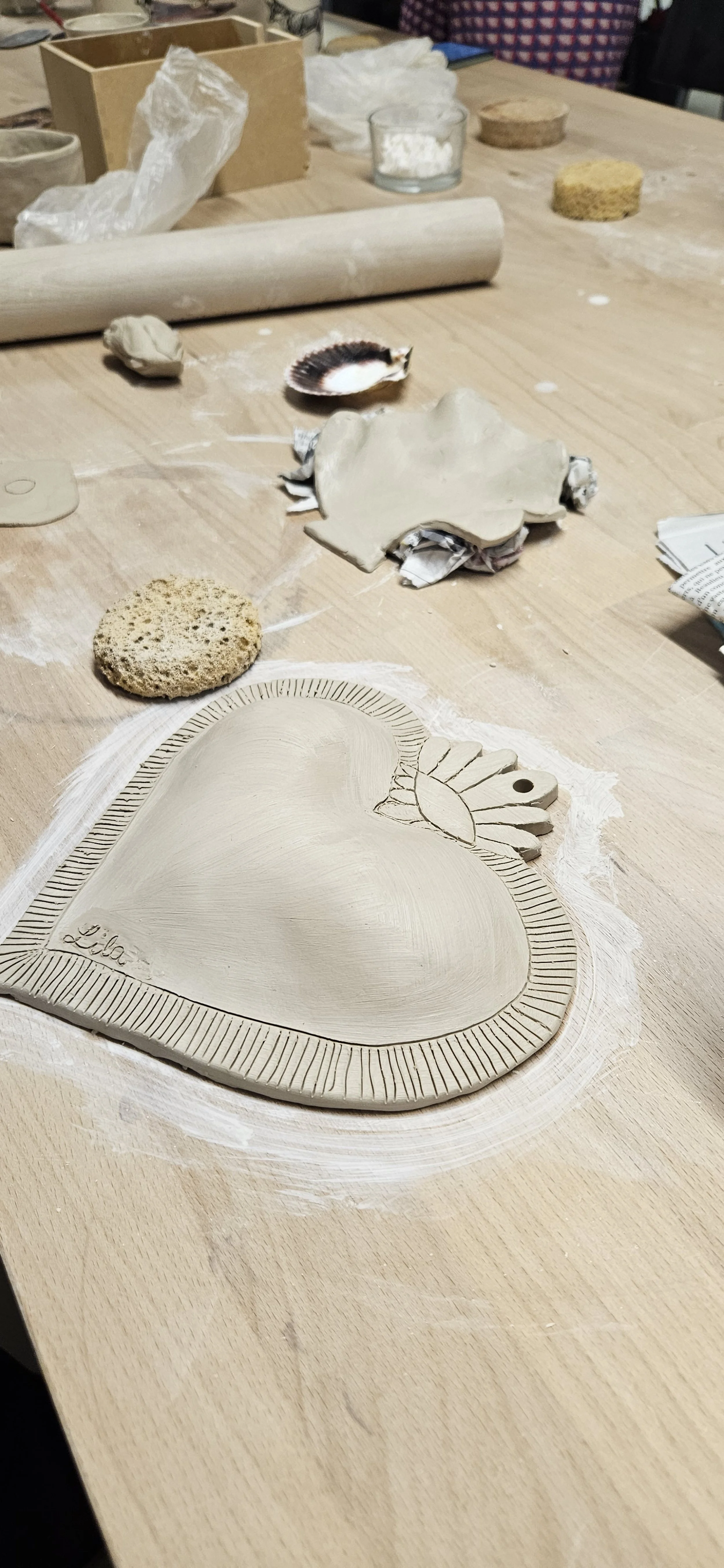 Une plaque en argile en forme de cœur avec des détails de soleil et des motifs gravés, portant le nom 'Lila', entourée d'outils et d'éponges pour la fabrication de céramique.