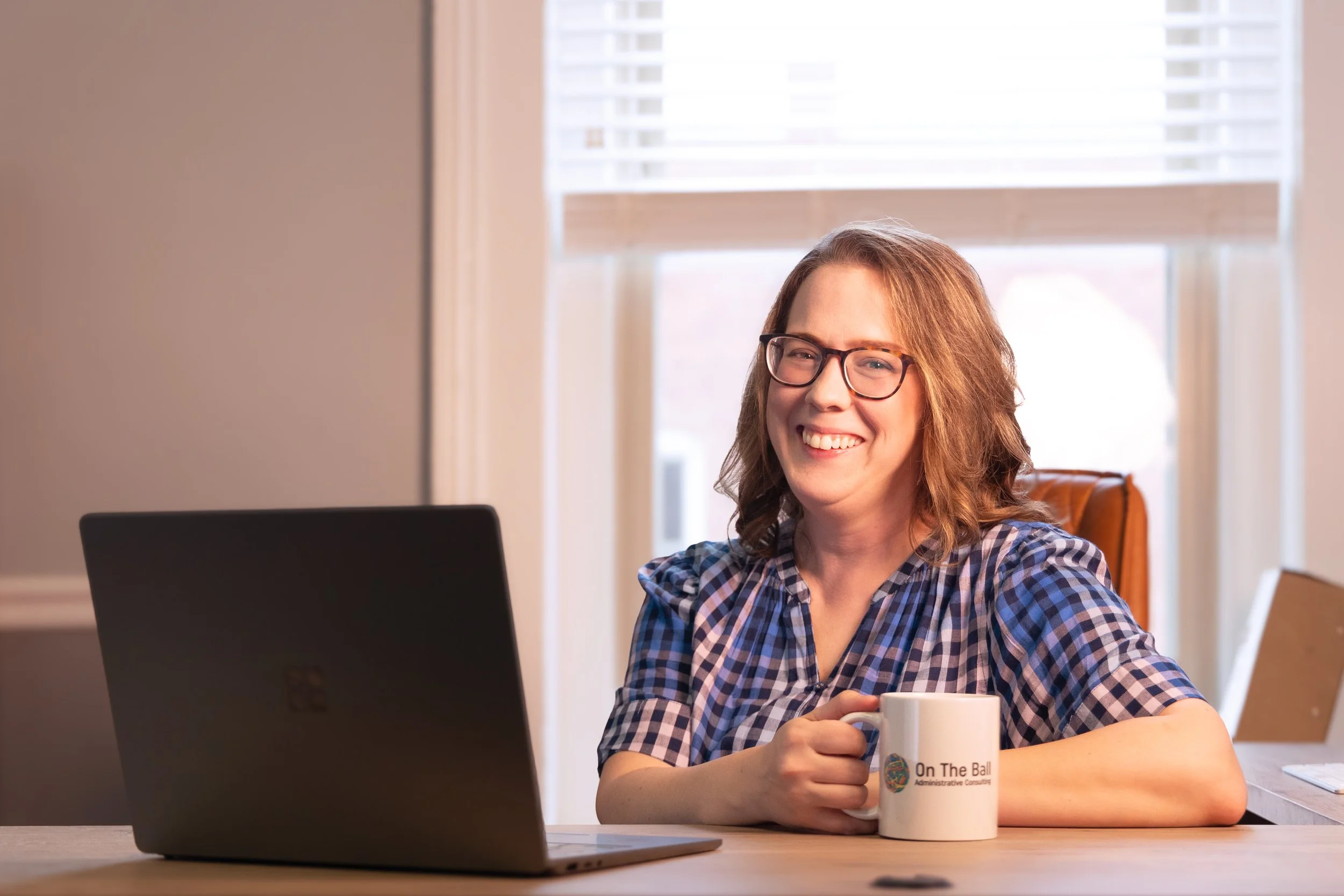Amanda Riley at her desk with her laptop in front of her and holding a coffee mug that says On The Ball