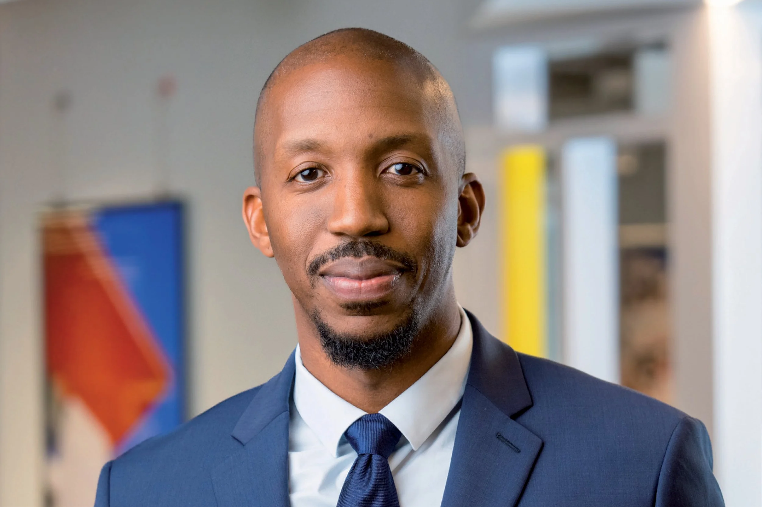 A man in a formal suit and tie smiling inside an office with a blurred American flag in the background.