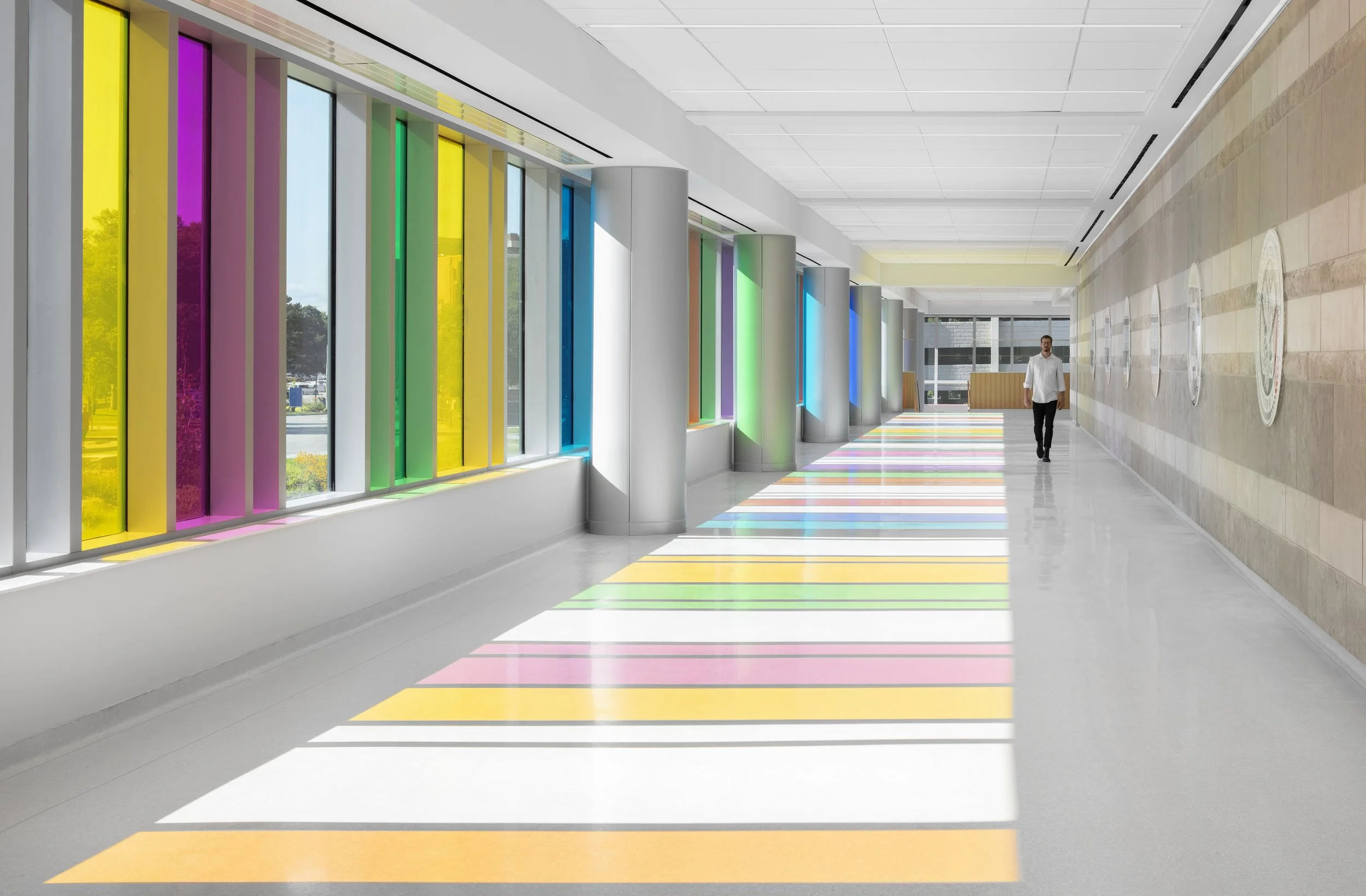 A man walks down a white hallway. To his left are large stained glass windows. The light is coming in through the window and casting colorful shadows on the floor.