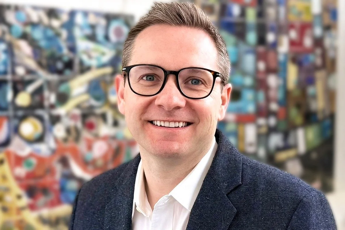 Portrait of Conner Holt, corporate controller, wearing a dark blazer and white shirt, standing in front of a colorful backdrop.