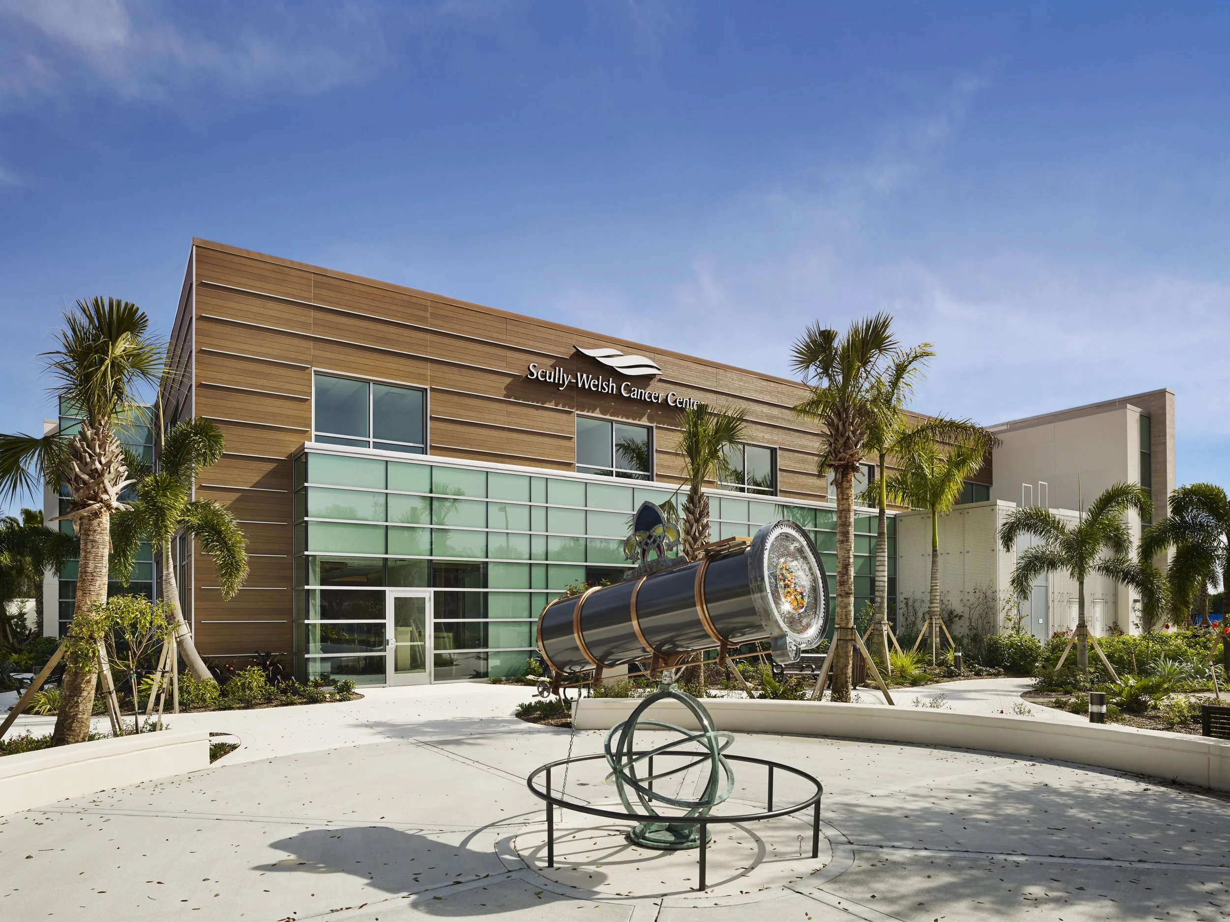 A large cylindrical object resembling a telescope sits on a decorative ball. A large building with a sign that says "Scully-Welch Cancer Center" is in the background surrounded by palm trees.