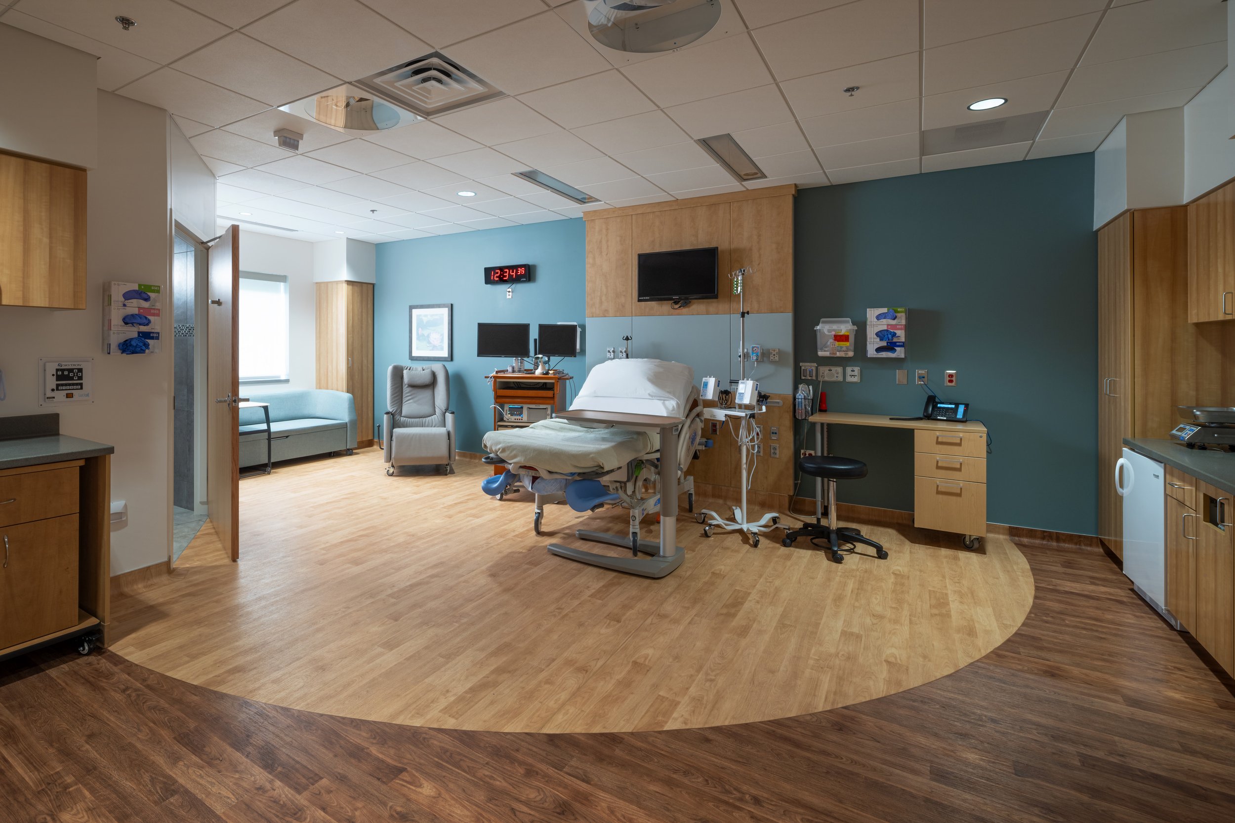 A large patient room with a patient bed, two tone wood floors, turquoise walls, medical equipment, a desk, a chair and couch, and a door to the bathroom.