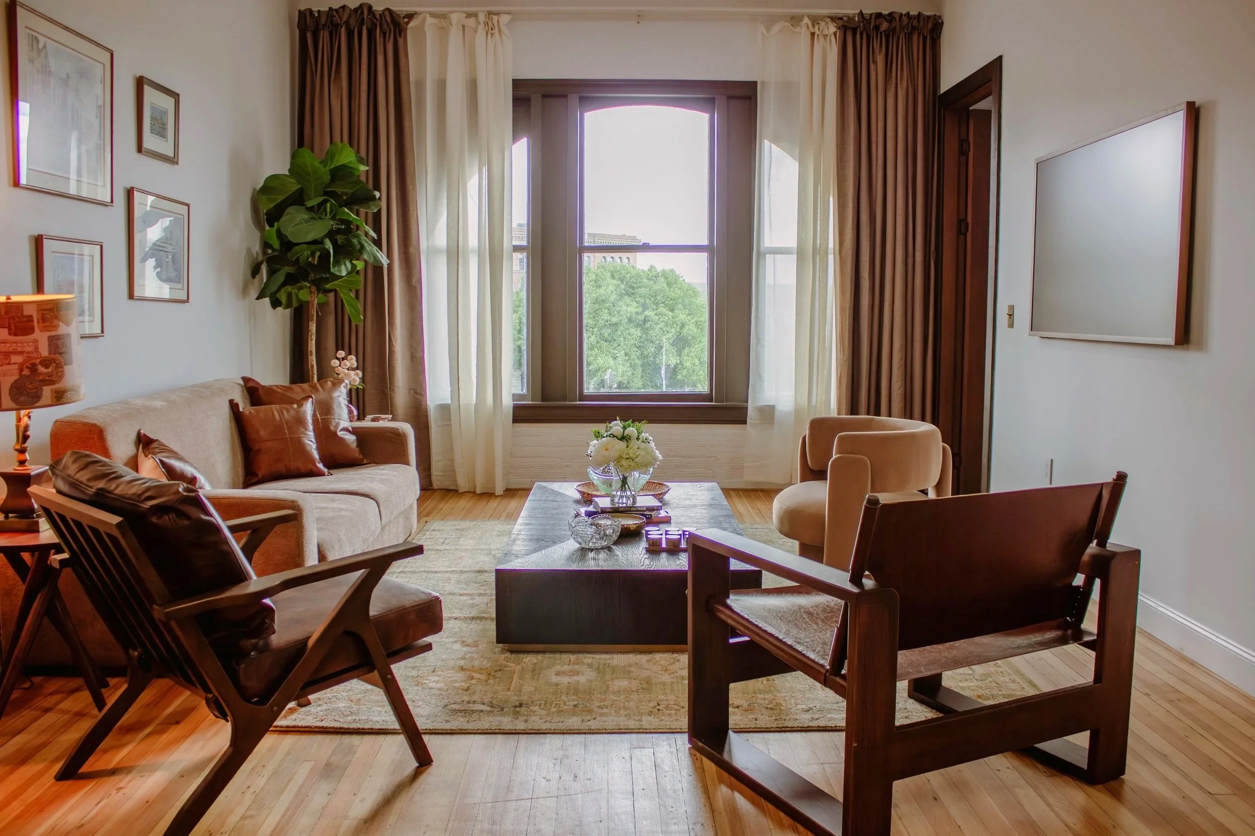 Living room with large window, beige sofa with brown pillows, wooden chairs, coffee table with flowers, framed art on white walls, large plant, and hardwood floors.