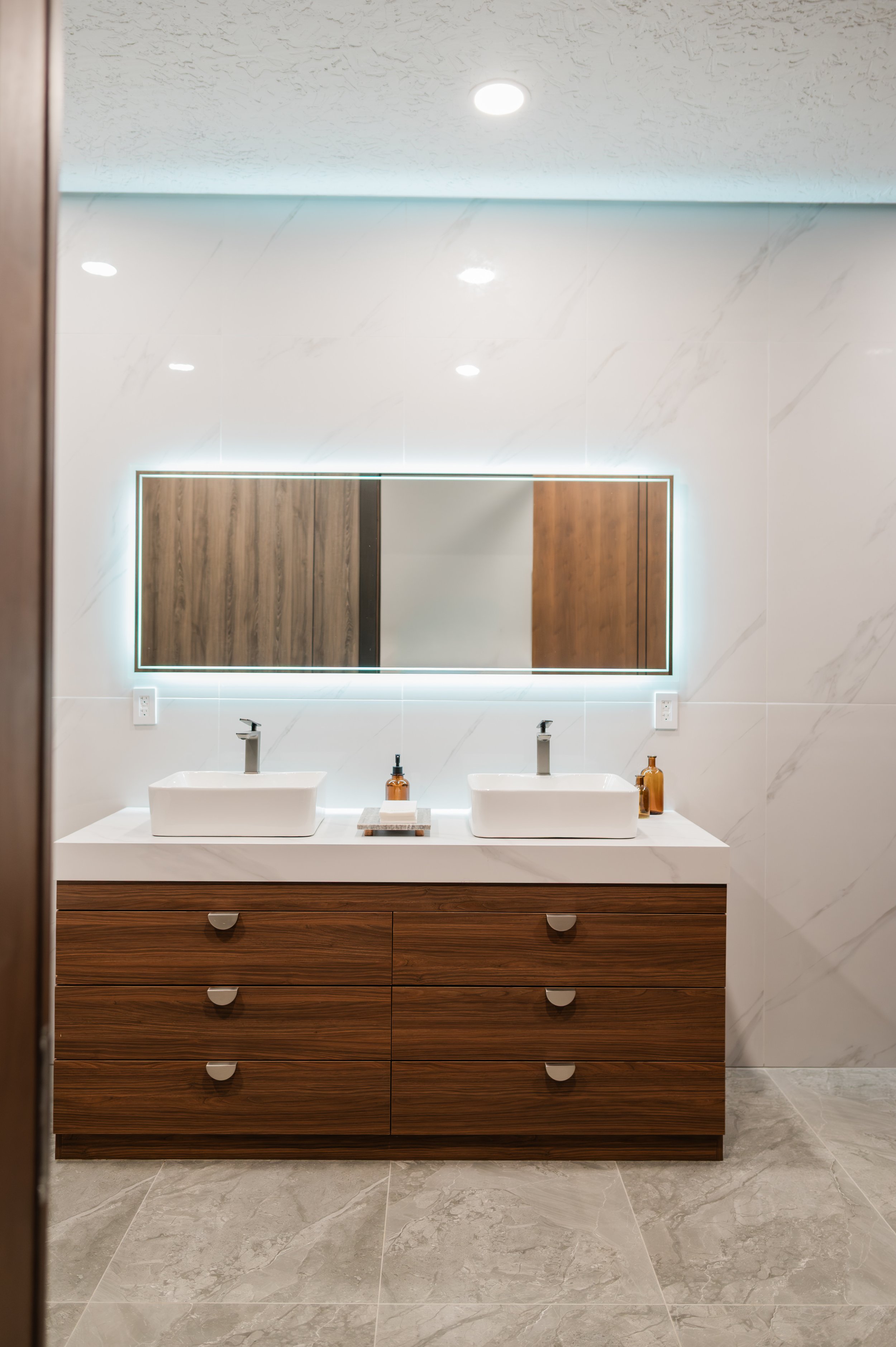 A modern bathroom with a large wooden vanity, two rectangular vessel sinks, and a large mirror with LED backlighting. The wall behind the mirror is covered in white marble tiles, and the bathroom has a textured ceiling with recessed lighting. Two amber bottles are placed on the vanity, and there are electrical outlets on either side of the mirror. The floor is covered with large gray marble tiles.