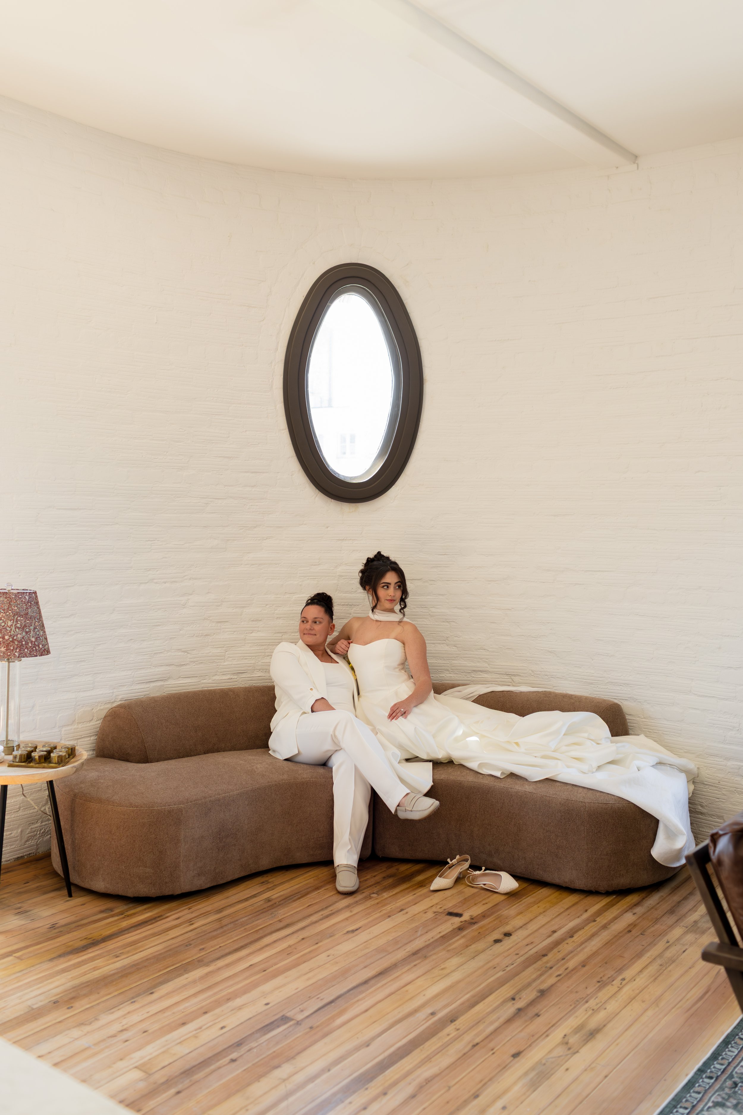 Two women sitting on a curved brown sofa in a room with a white brick wall and a small, oval window. One woman is wearing a white suit and the other is in a white wedding dress, with their shoes on the floor in front of them. There is a lamp with a floral shade on a side table to the left.