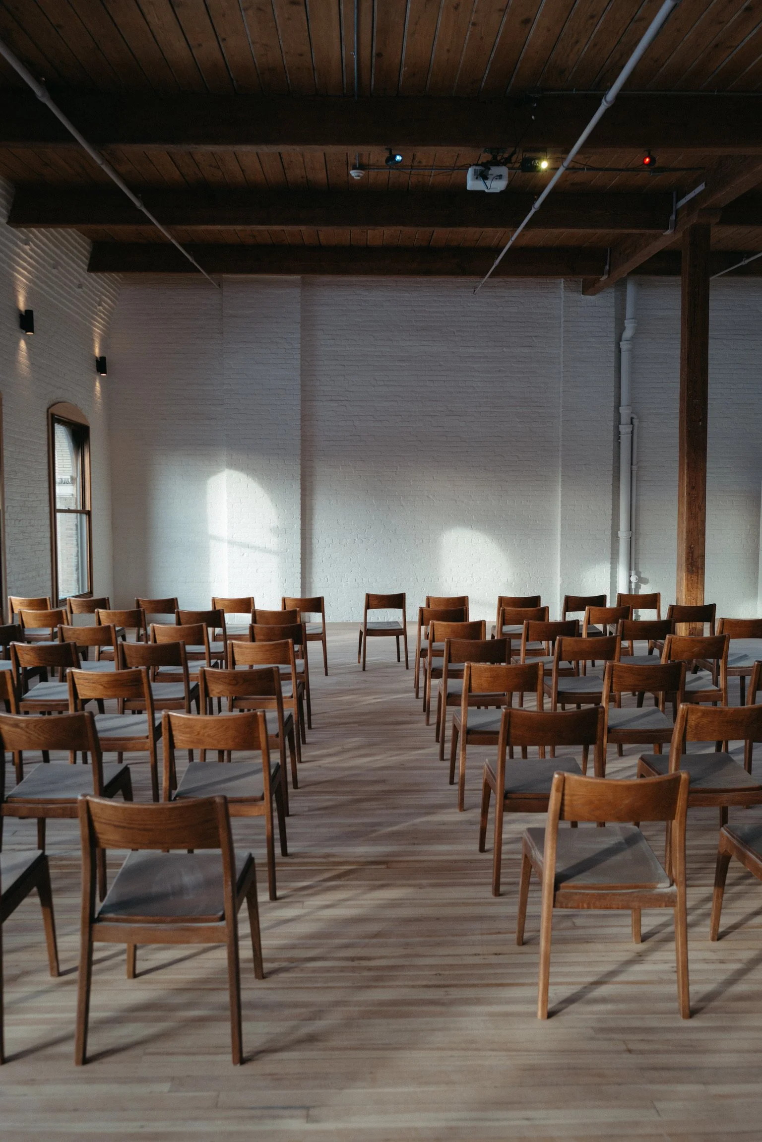 Empty wooden chairs arranged in rows in a spacious room with white brick walls, large windows, and a wooden ceiling with exposed beams.