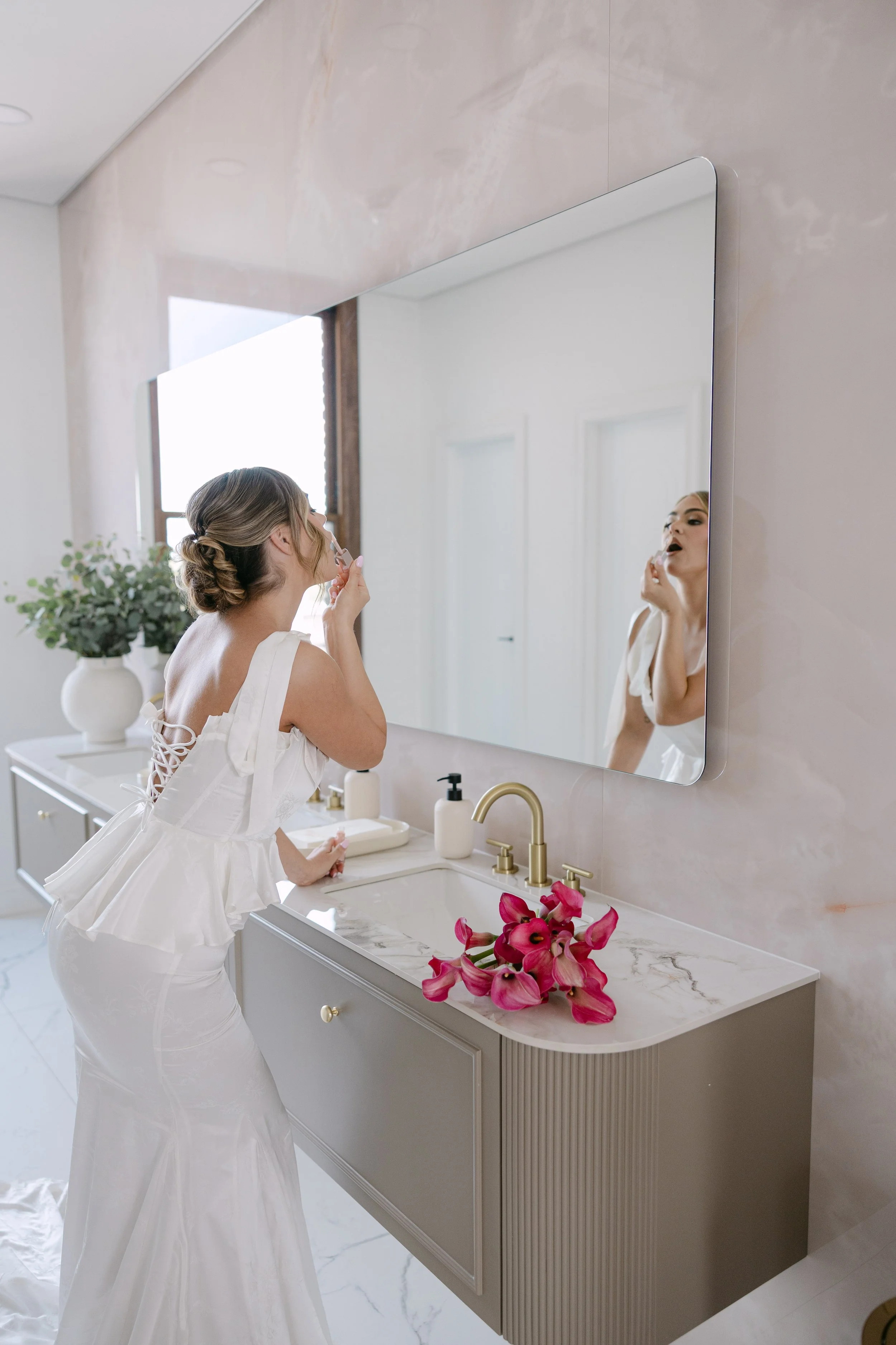 A woman in a white dress is applying lipstick in front of a large bathroom mirror. The bathroom has a light-colored marble countertop with a pink flower arrangement and soap dispensers. A potted plant is visible on a cabinet behind her.