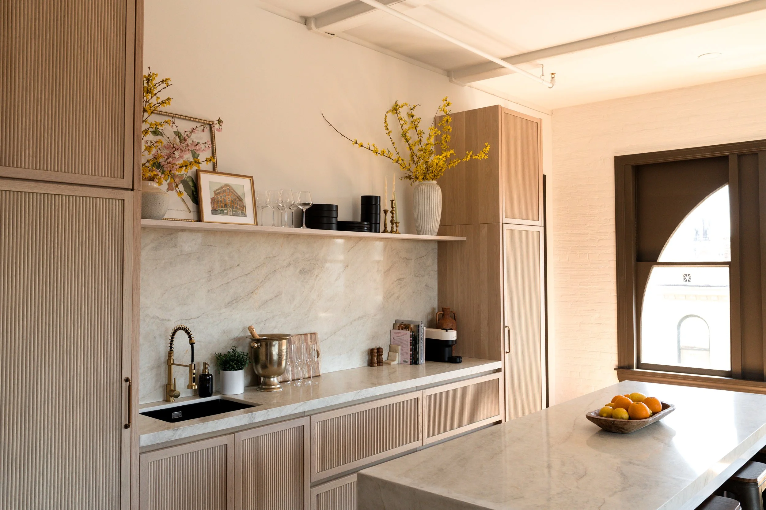 A modern kitchen with light wood cabinets, a white marble countertop, and a large window. Decorative items include a framed picture, glassware, vases with flowers, and a bowl of oranges on the island.