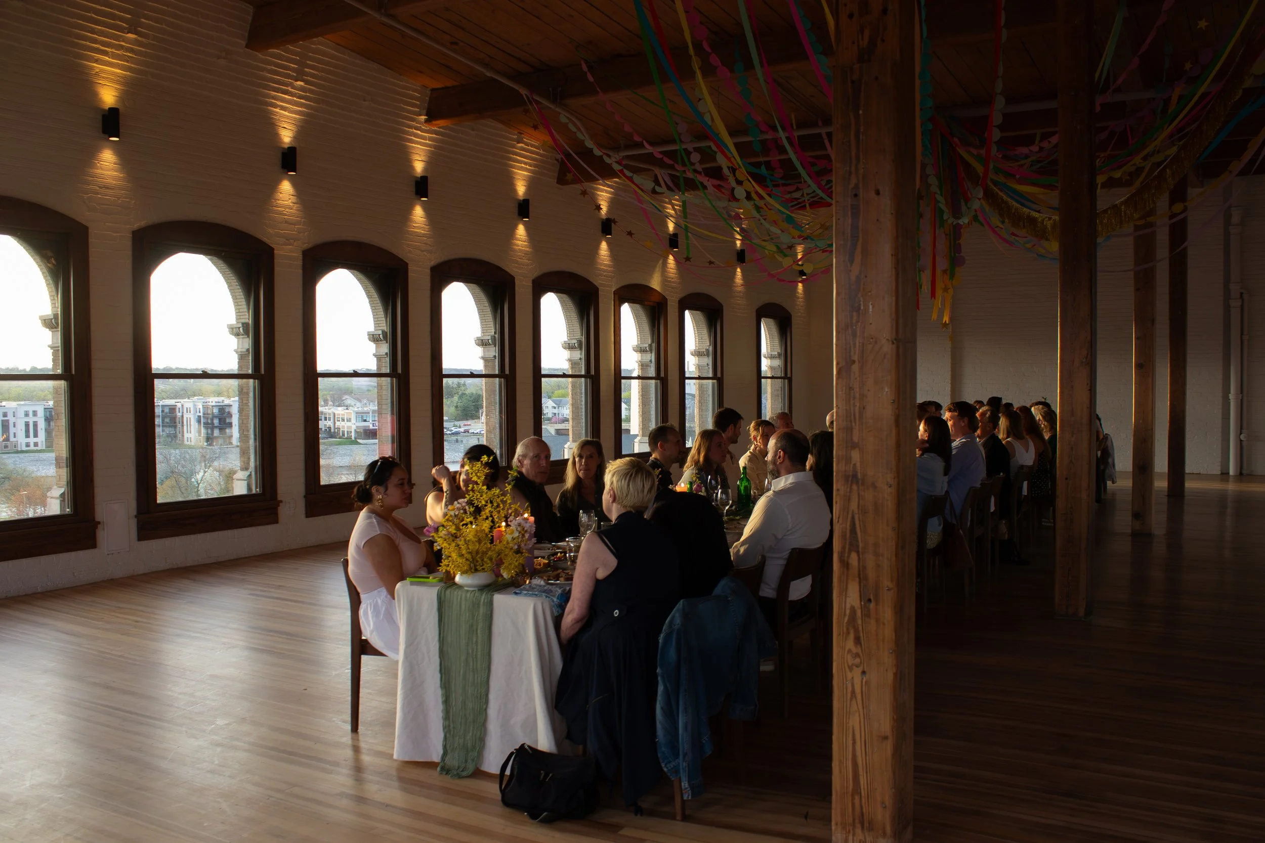 People sitting at a decorated banquet table during a celebration or event, with large windows and wooden beams in the room.