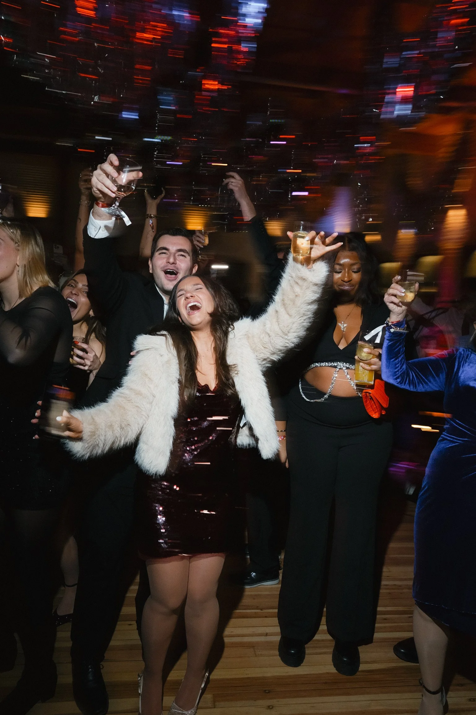 People celebrating at a party, holding glasses of drinks, wearing formal and festive attire, with colorful lights and blurred motion effects in the background.