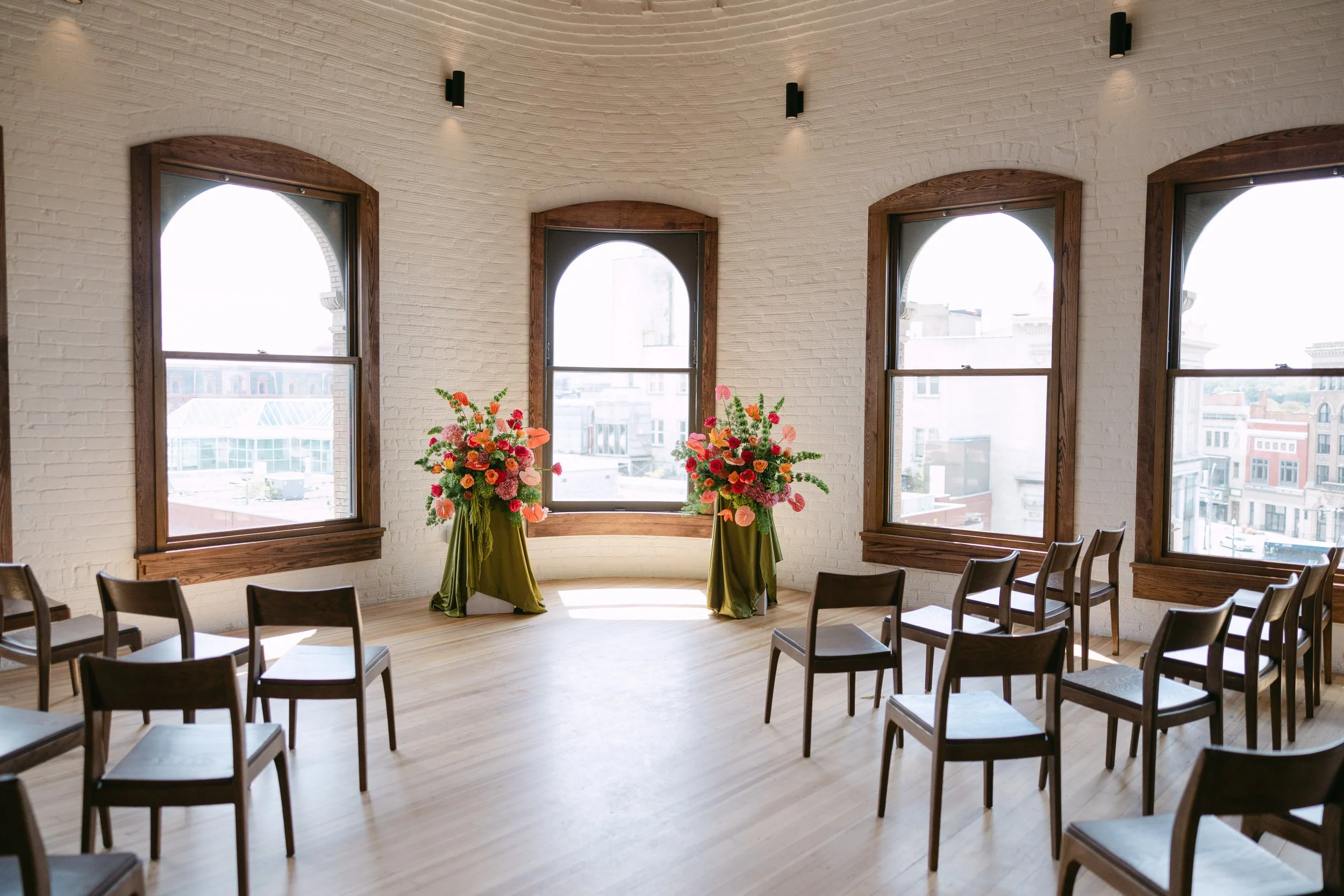 An empty indoor room decorated with two floral arrangements on tall stands and surrounded by chairs, with large arched windows allowing natural light to fill the space.