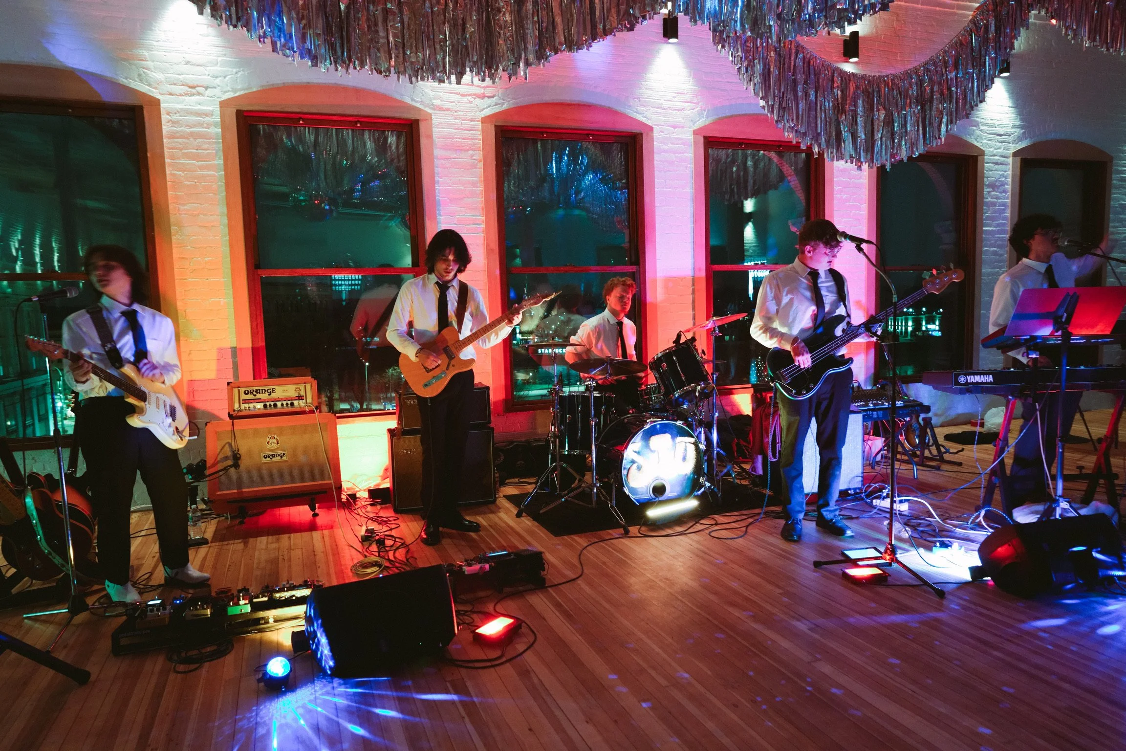 A band performing on stage with guitars, drums, keyboard, and microphones in a venue decorated with hanging multicolored tinsel and blue and purple lighting.