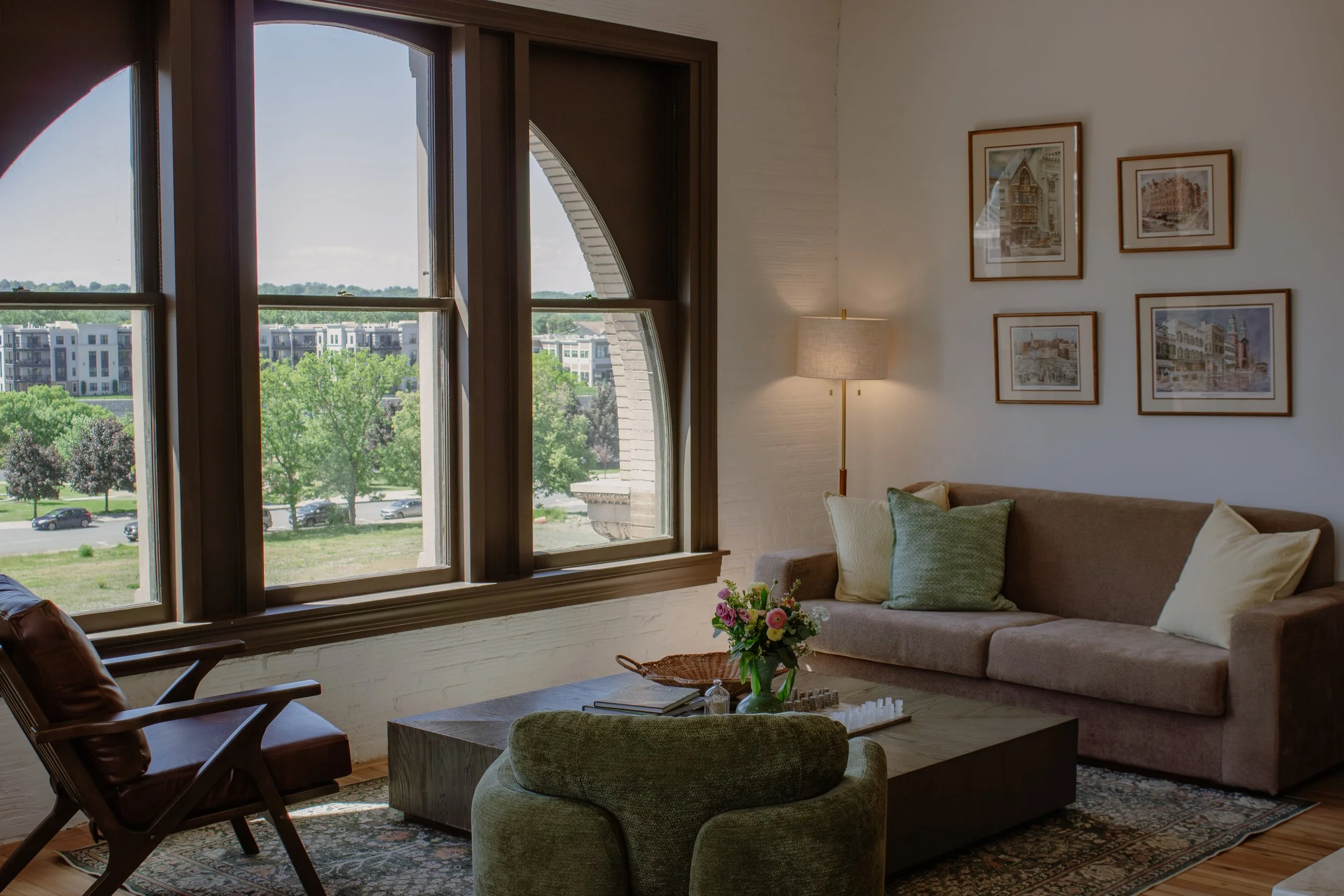Living room with large windows overlooking a green outdoor area with modern white buildings, decorated with framed pictures on the wall, a floor lamp, and a sectional sofa with pillows.