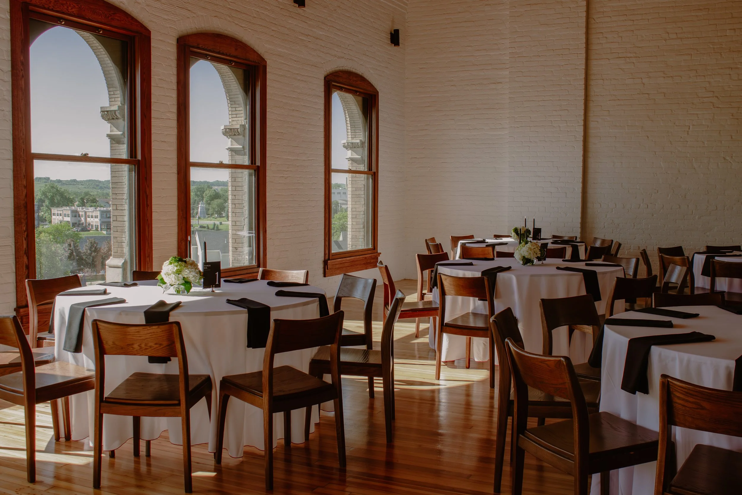 Interior of a restaurant with white brick walls, large arched windows, and wooden chairs around round tables with white tablecloths and black napkins.