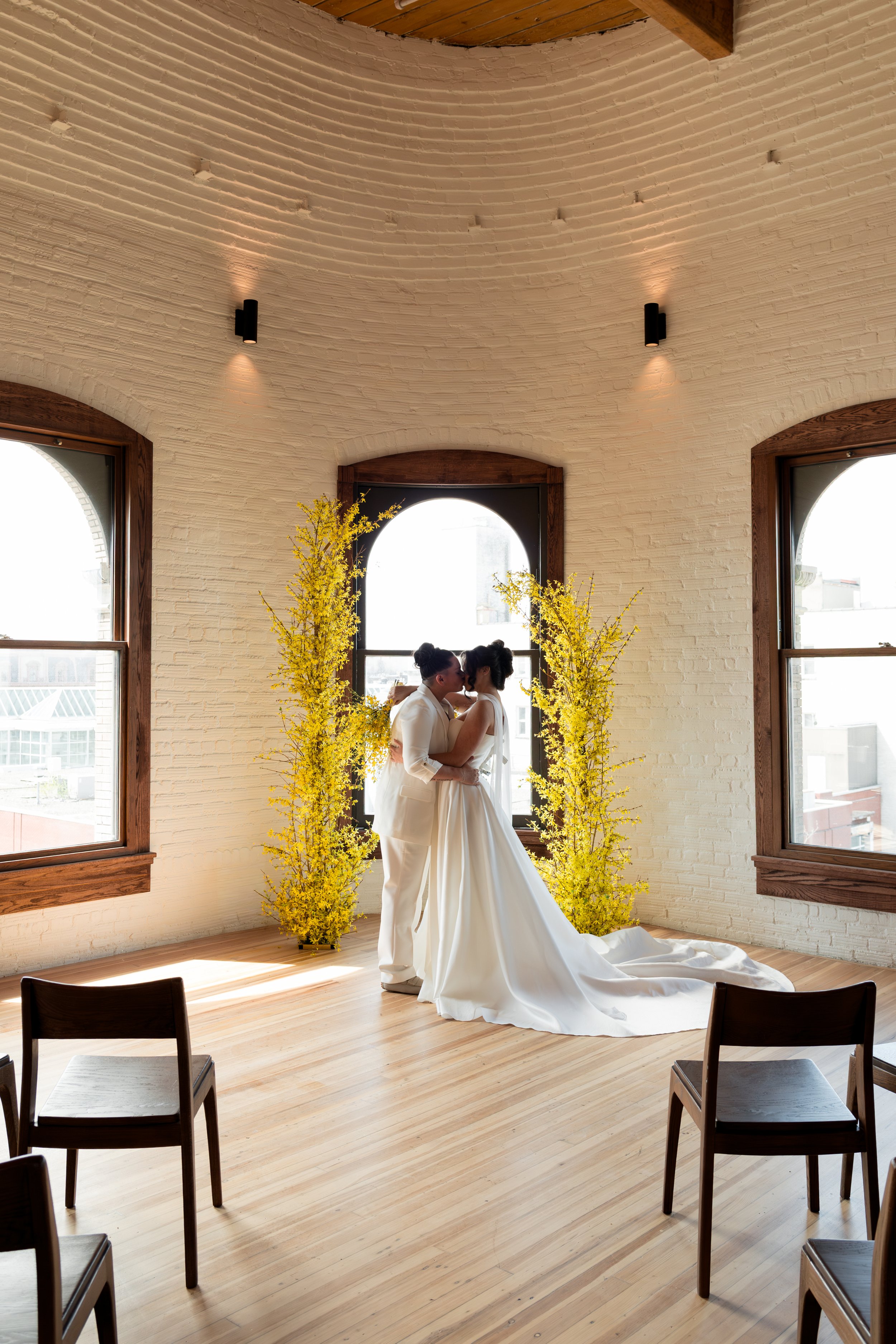 Couple in wedding attire sharing a kiss in a bright, airy room with large windows, wooden floors, and decorative yellow trees.