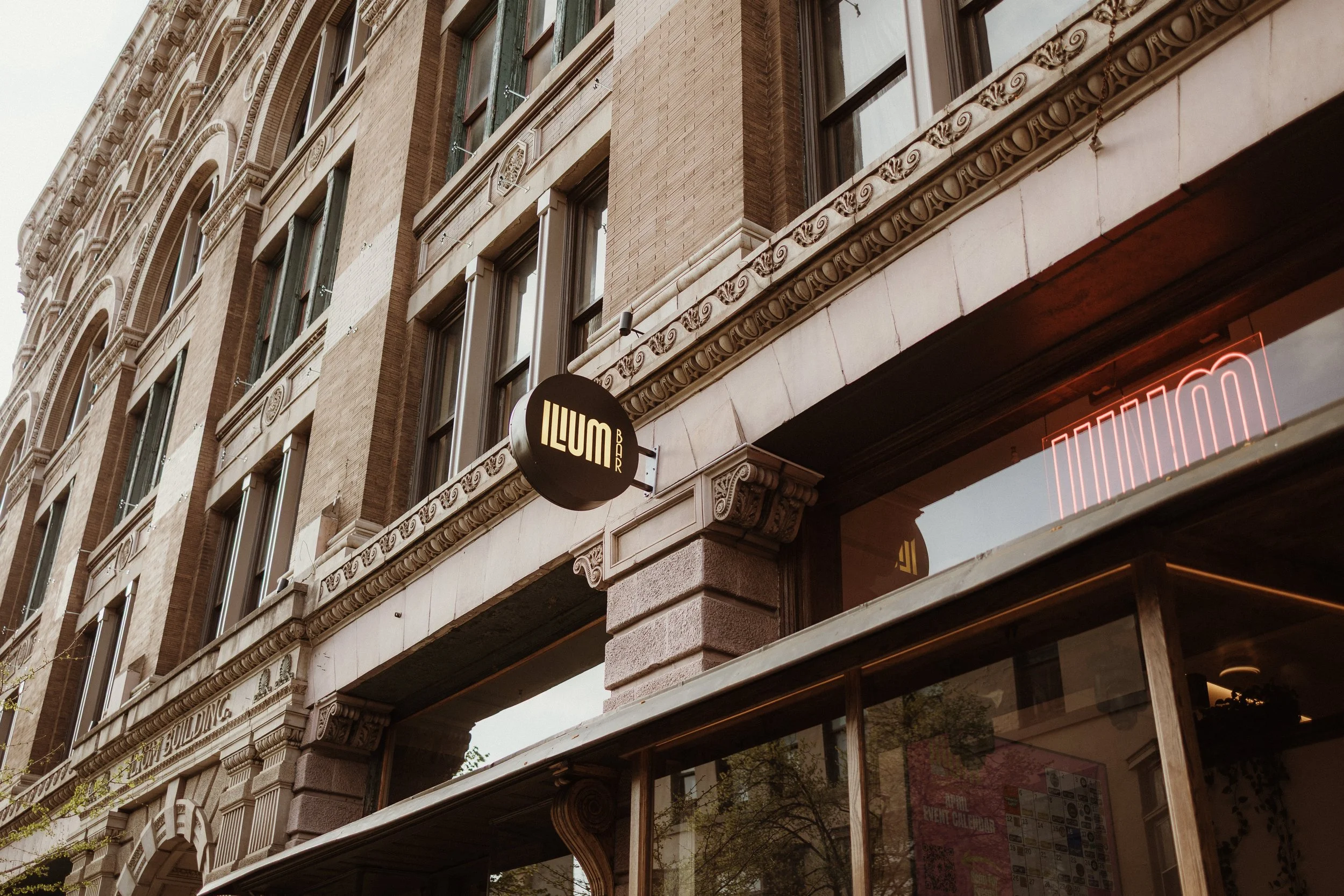 Close-up of a historic multi-story brick building with ornate architectural details, large windows, and a lit neon sign that reads 'ILLUM BAR'.