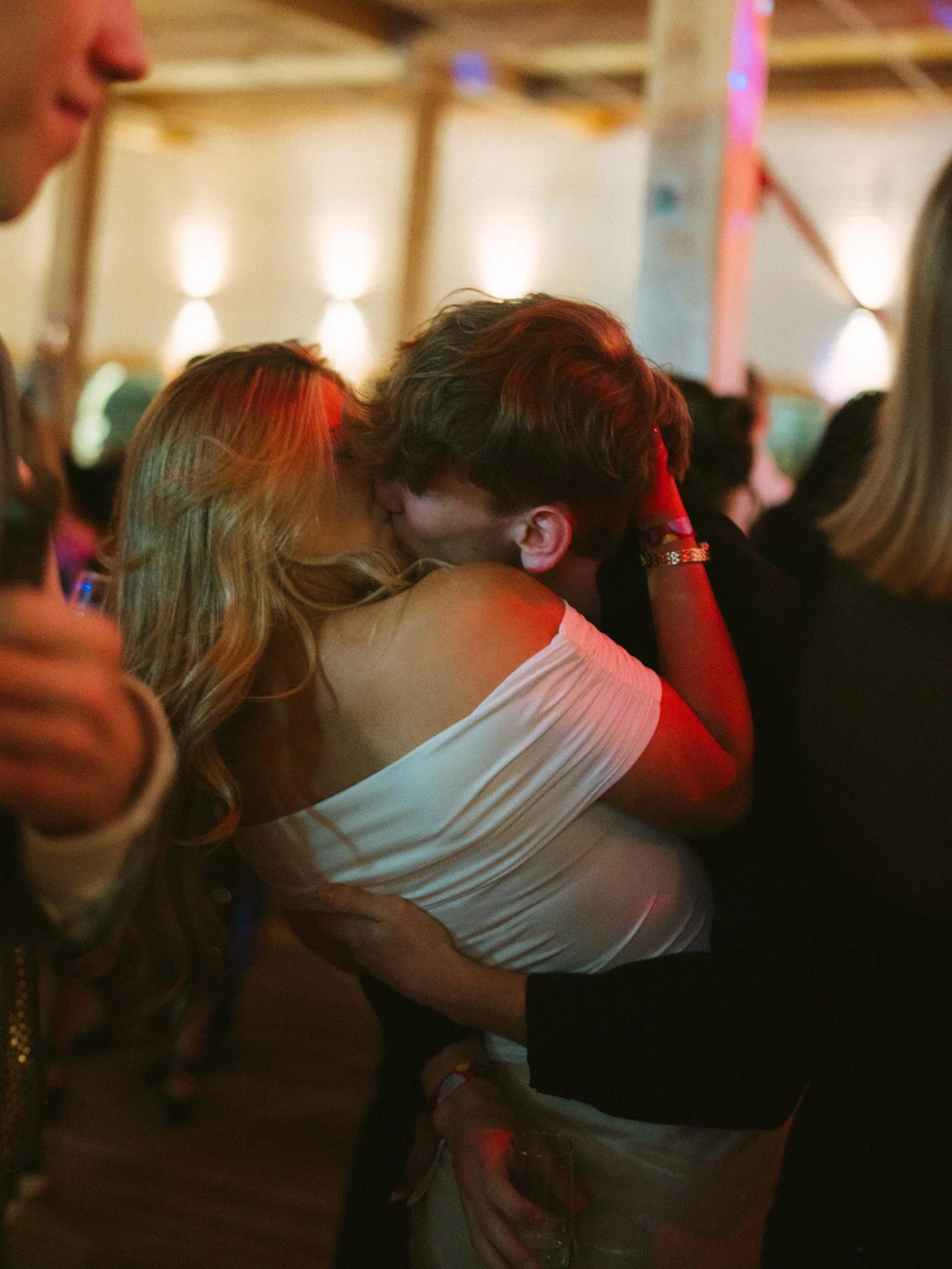 Two people kissing at a social event or party. The scene is indoors with warm lighting and blurred background, showing other people socializing.