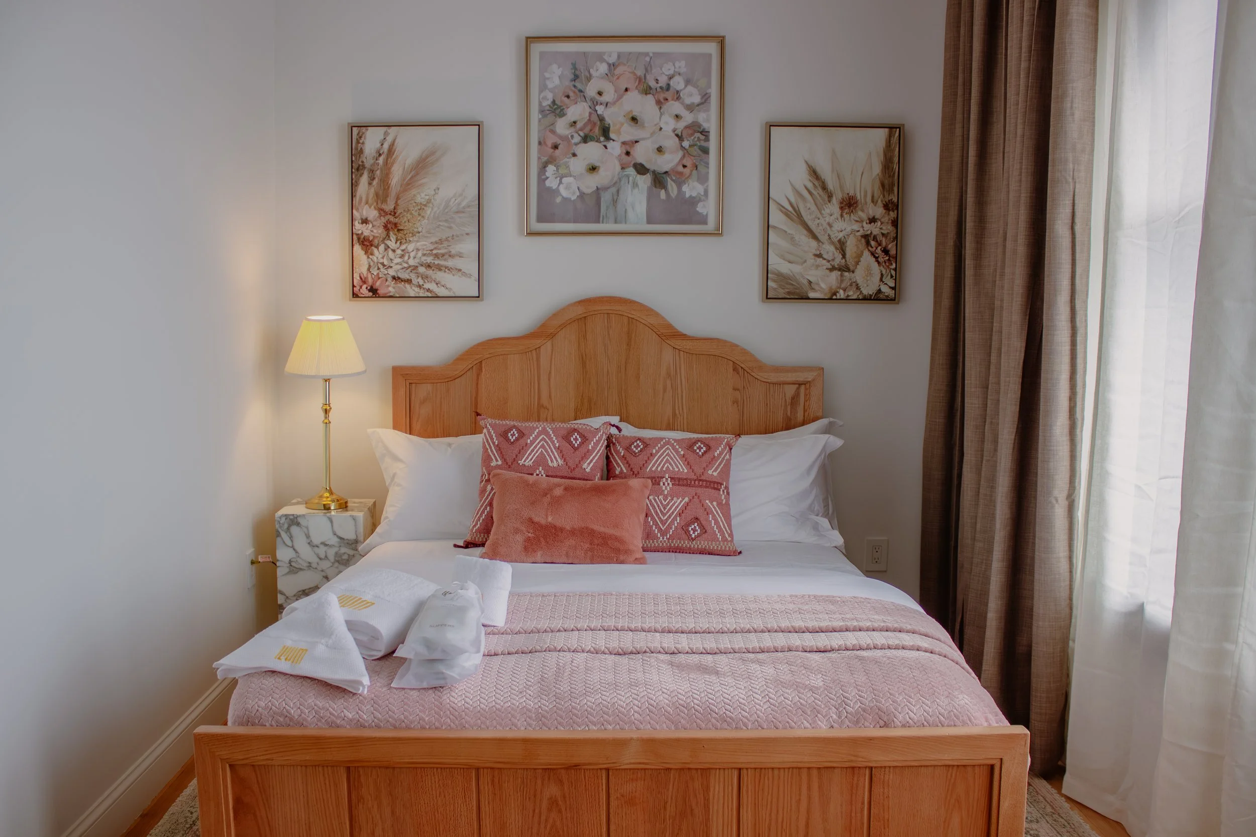 A neatly made bed with pink and coral pillows, a pink blanket, and white sheets, a marble nightstand with a lamp, and framed floral artwork on the wall, in a cozy bedroom with curtains.