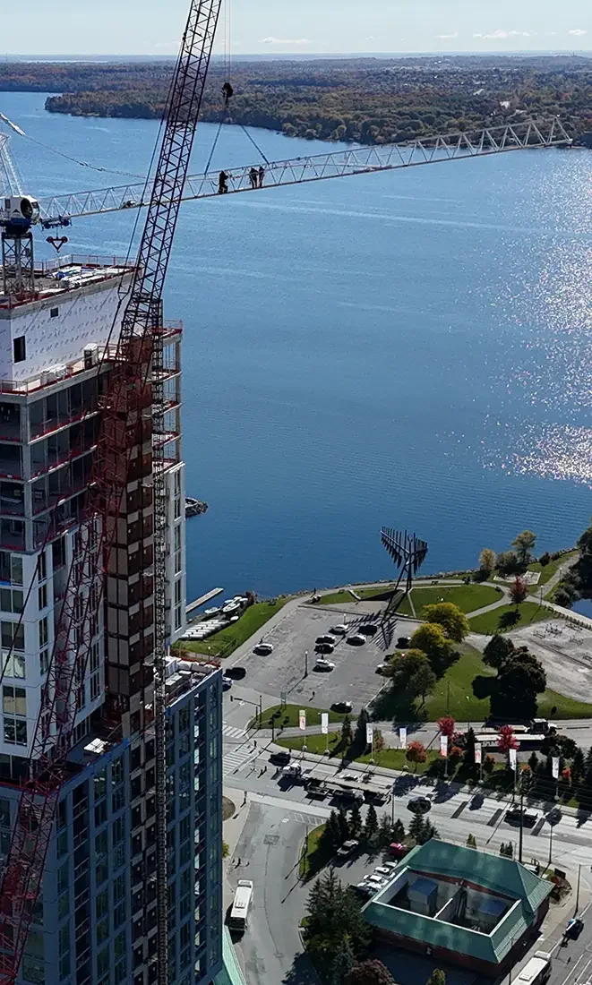 View of a cityscape in Barrie, ON with a body of water, a large construction crane installing a bridge, and a park with an antenna structure in the foreground.