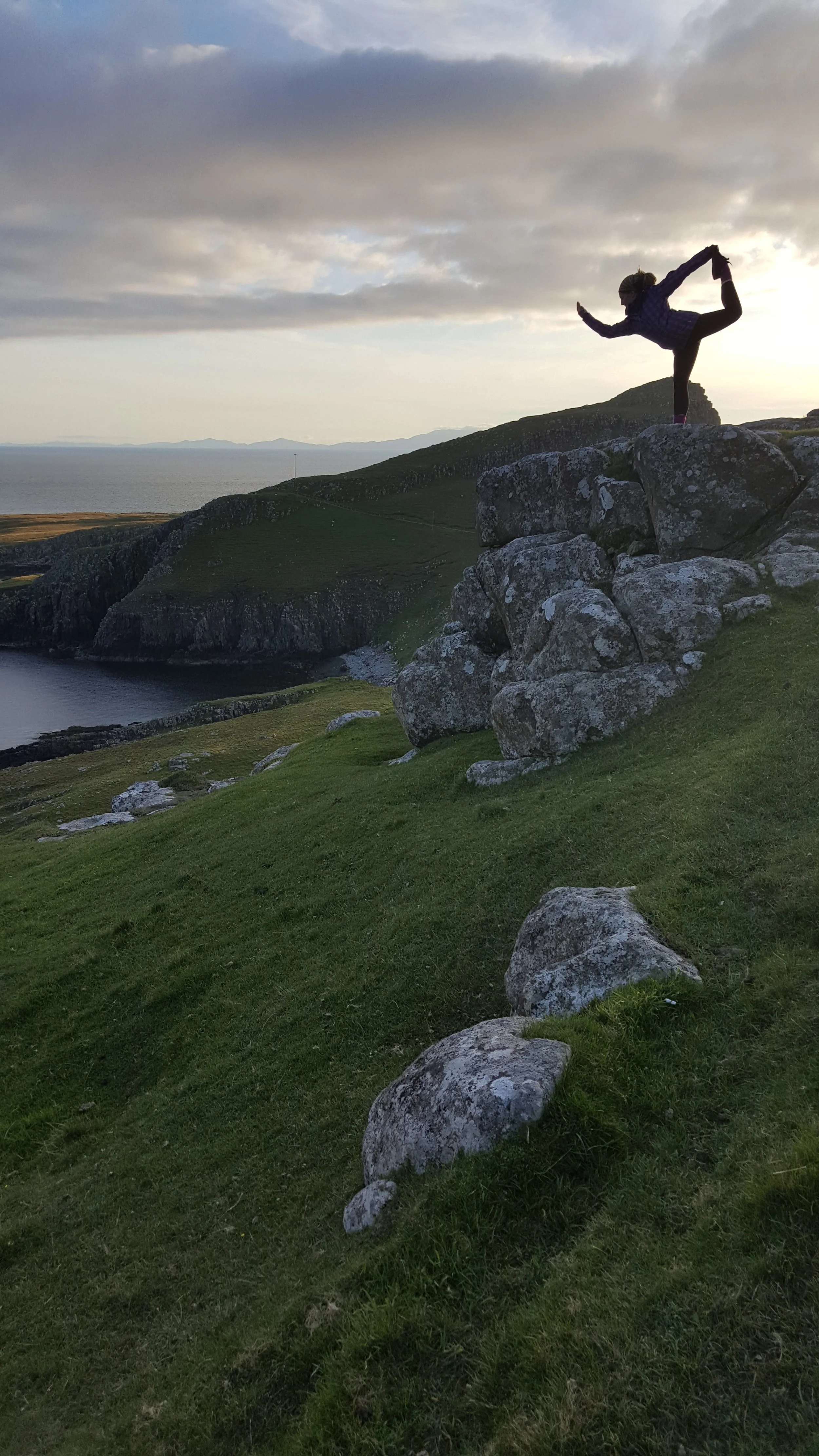 A person standing on rocks outdoors, balancing in a yoga pose, with a scenic view of rolling green hills, water, and a cloudy sky in the background.