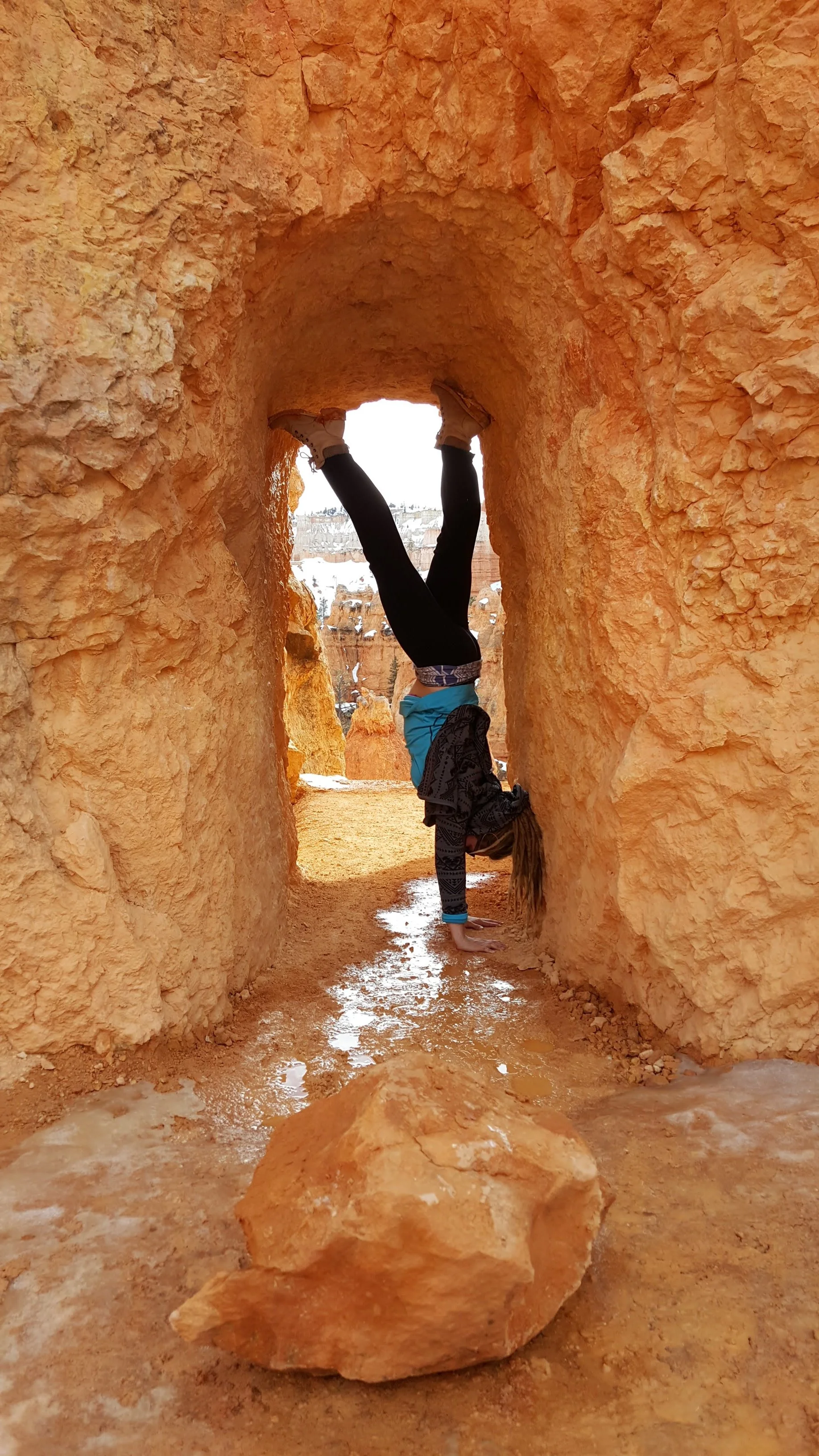A person is performing a handstand against a red rock formation with a natural arch, in a desert landscape, with snow visible in the background.