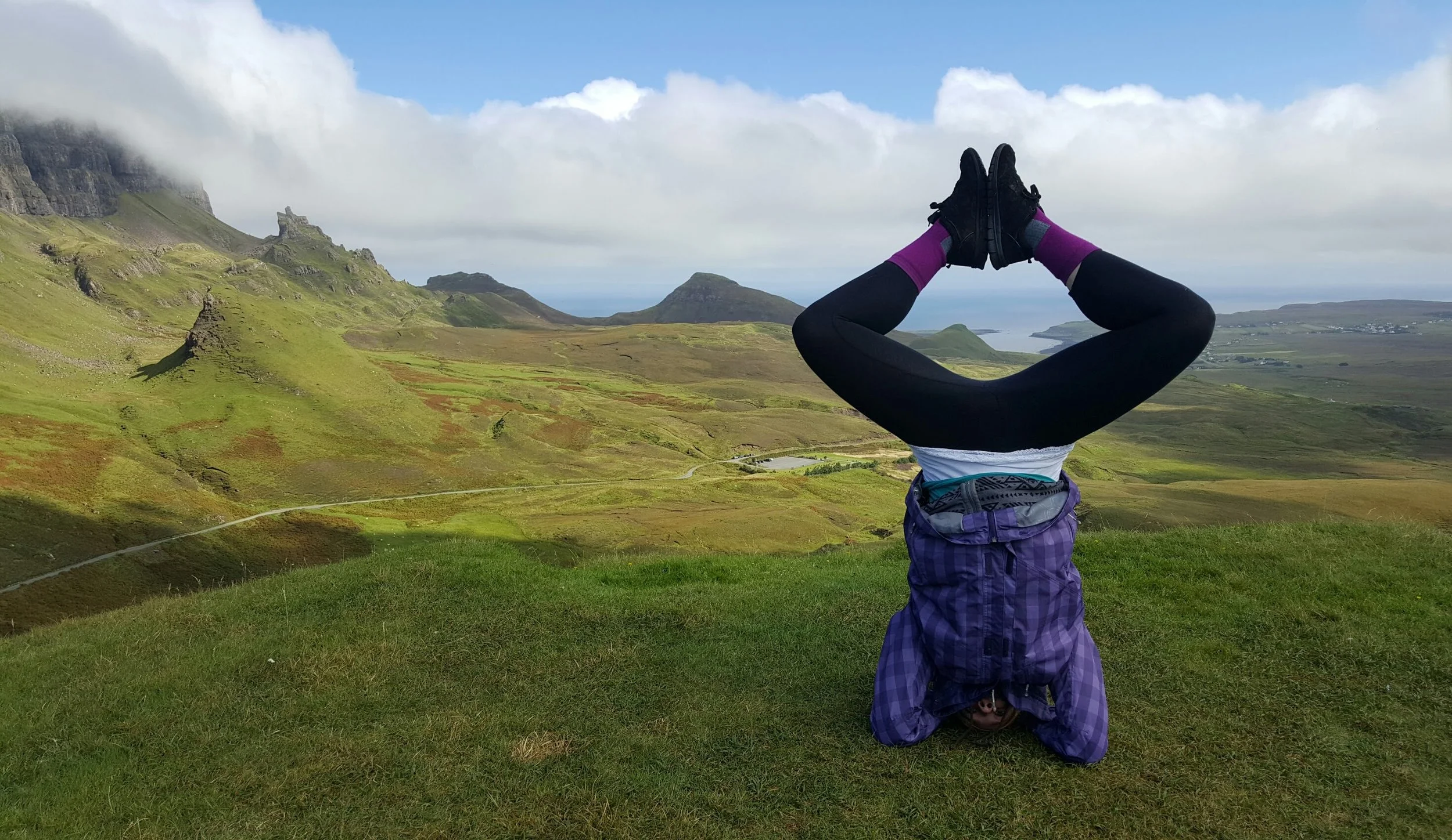 A person standing upside down with their head on the ground and legs in the air, facing a scenic landscape of green hills, mountains, and water, during daytime with partly cloudy sky.