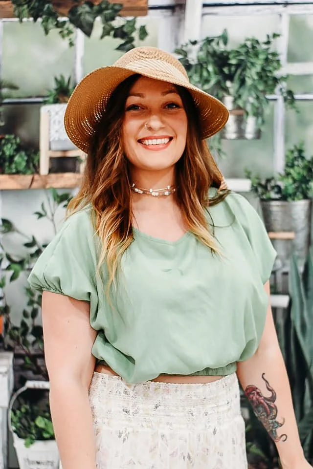 A woman with long, wavy hair wearing a straw hat, a light green crop top, and a white skirt, smiling in what appears to be a greenhouse or plant shop with various potted plants in the background.