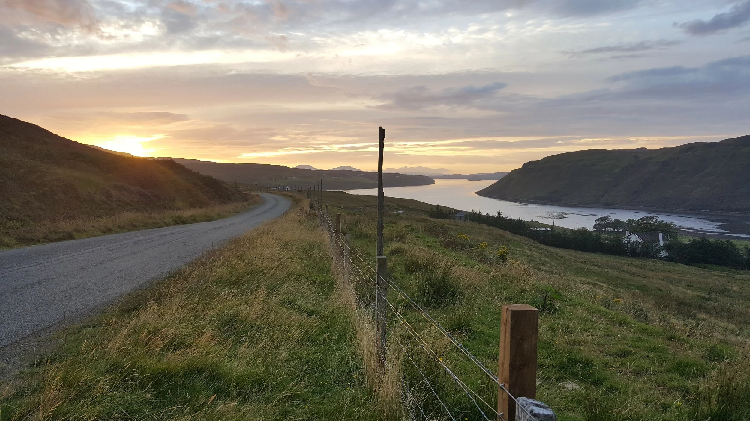 Scenic landscape of a winding road through grassy hills with a view of a river and distant mountains during sunset, with scattered clouds in the sky.