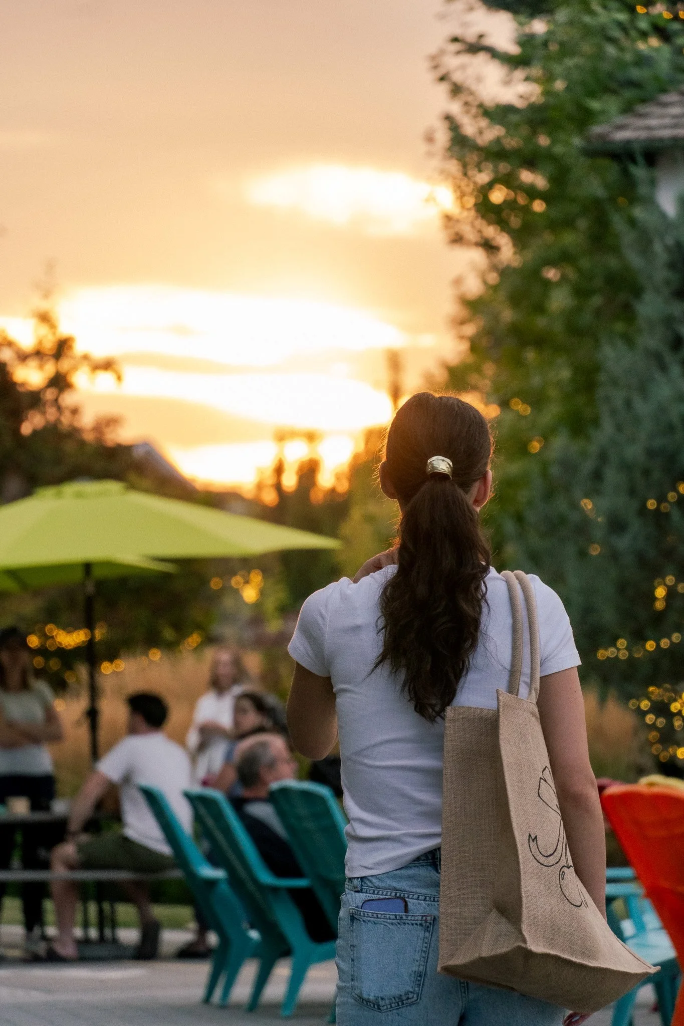 Rear view of a woman with a tote bag watching a golden hour sunset at a community event captured by Aces and Oak.