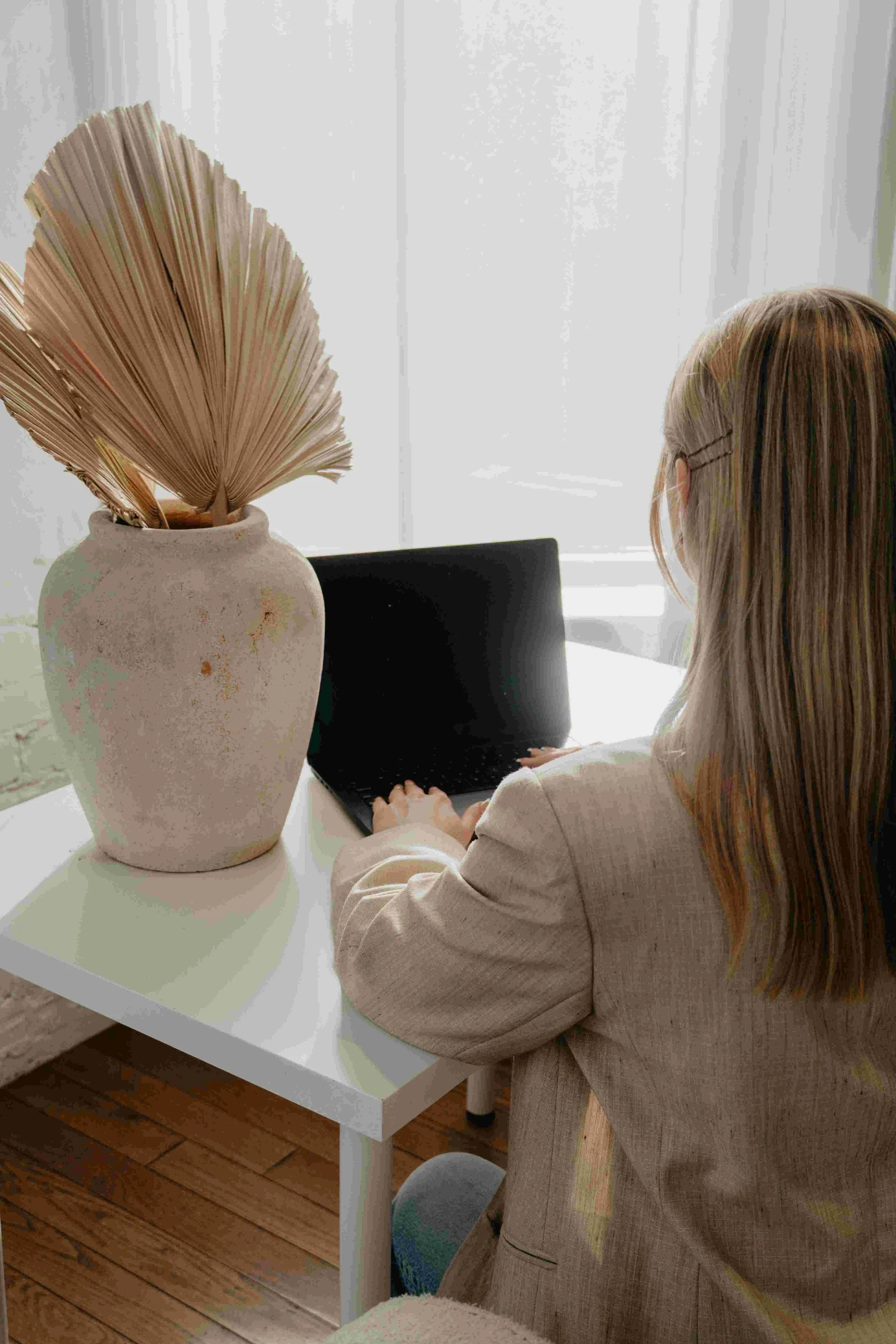 A minimalist white desk featuring a ceramic vase with dried fan palm leaves in a brightly lit studio.