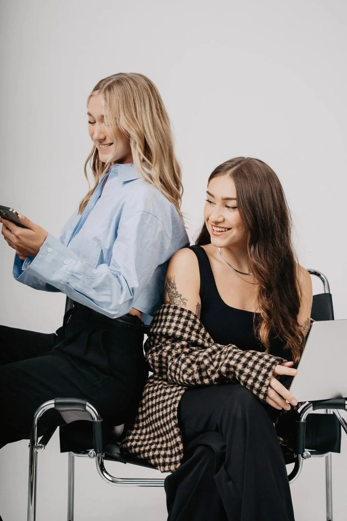 Two smiling agency owners sitting back-to-back using a smartphone and laptop during a brand photo shoot by Aces and Oak.