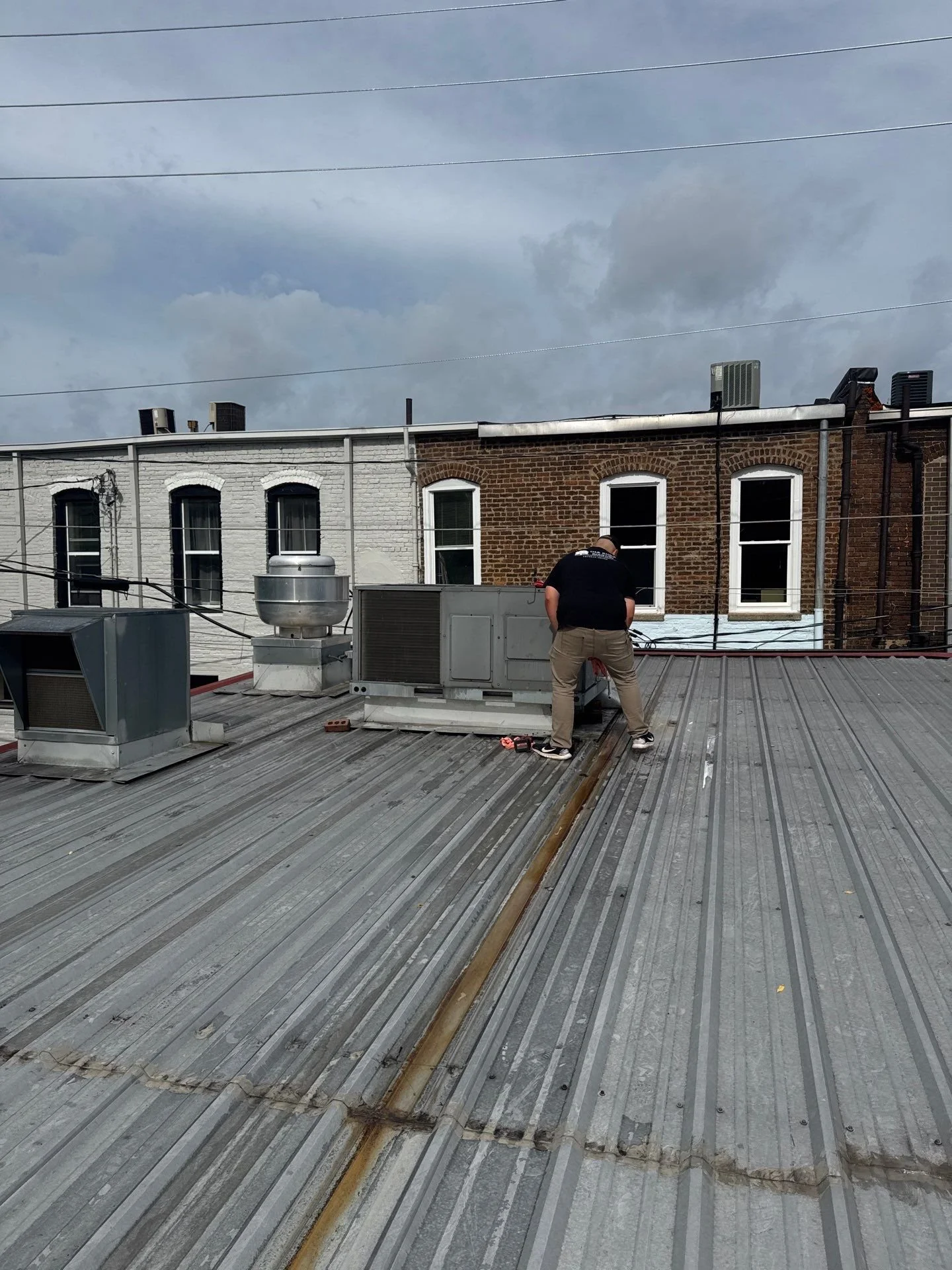 A man working on a rooftop with HVAC units and vents, with row of brick buildings with white and brown brick facades, and stormy sky in the background.