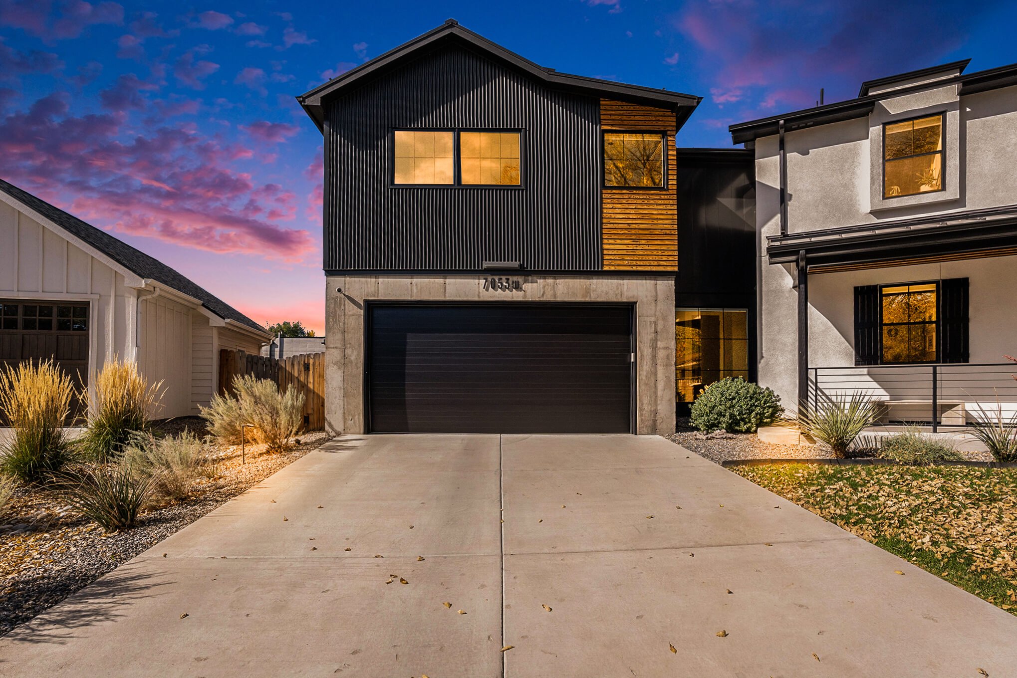 Modern black and white two-story house with a garage, minimalist landscaping, and sunset sky.