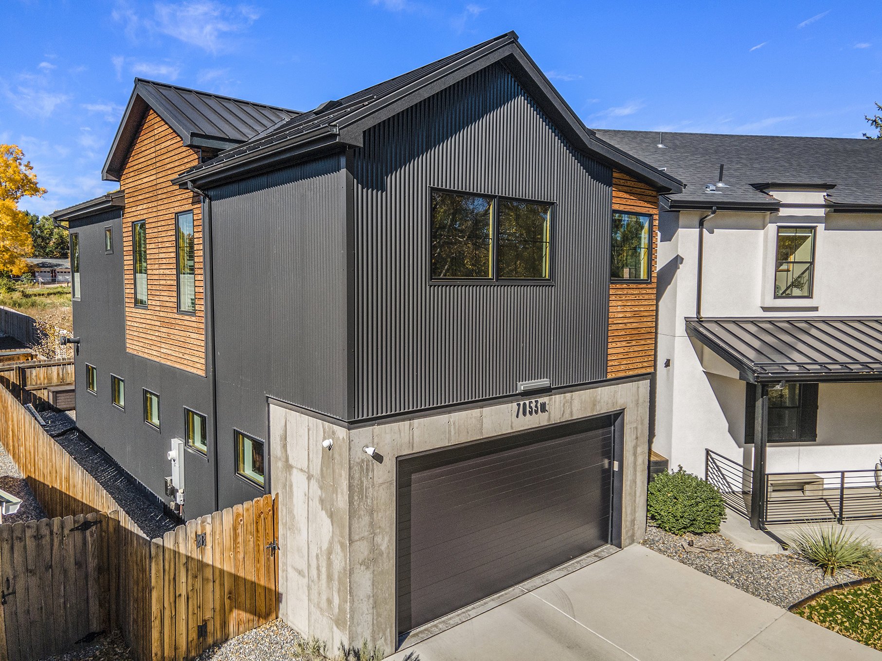 Modern-style house with a combination of wood and black metal siding, featuring multiple windows and a garage. The house is adjacent to a white building with a metal roof and surrounded by a wooden fence.