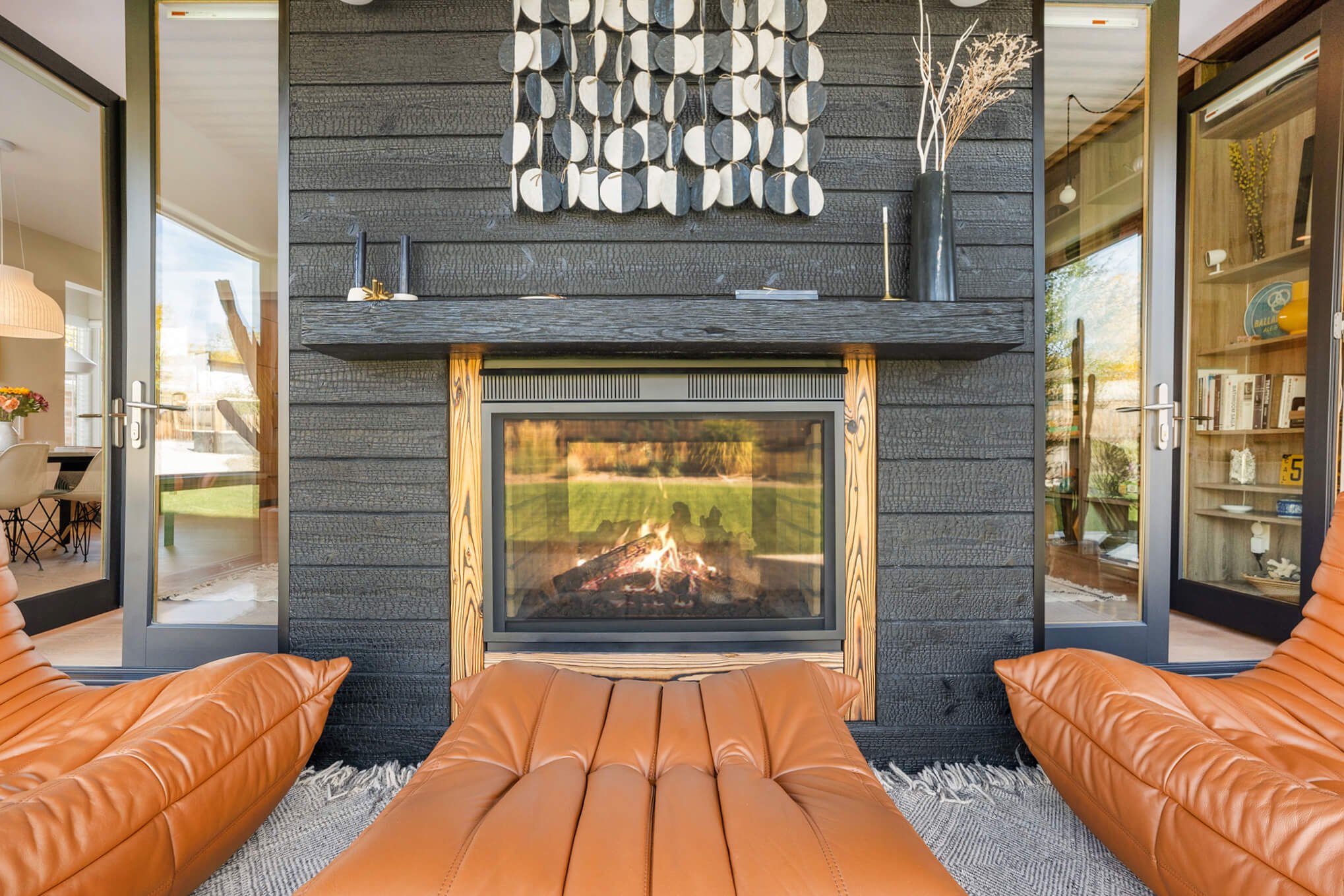 Contemporary outdoor fireplace with orange leather lounge chairs and modern decor accents, surrounded by glass doors.
