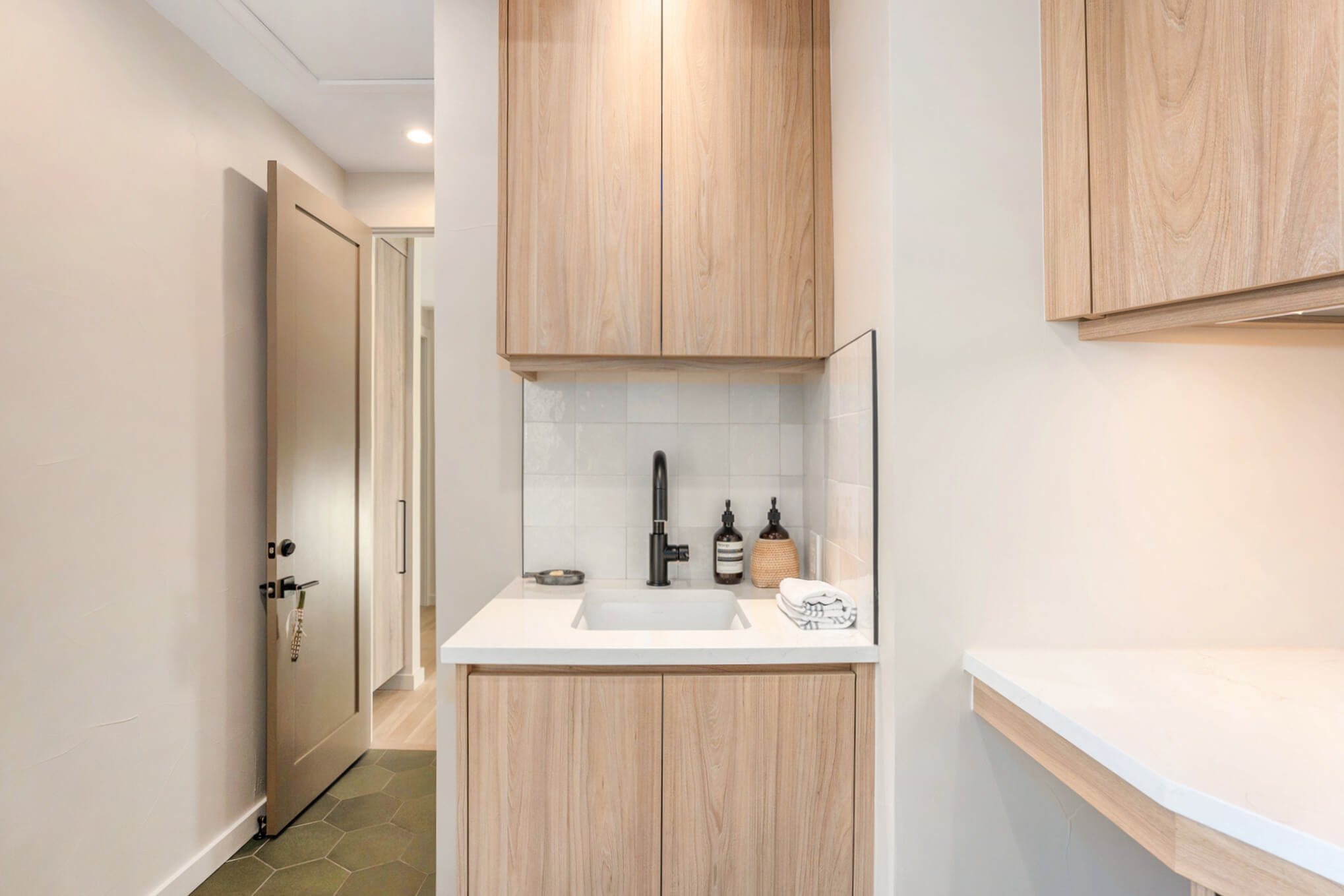 Modern laundry room with wooden cabinets, black faucet, and tile backsplash above a sink, featuring soap dispensers and folded towel, with an open door leading to an adjacent space.