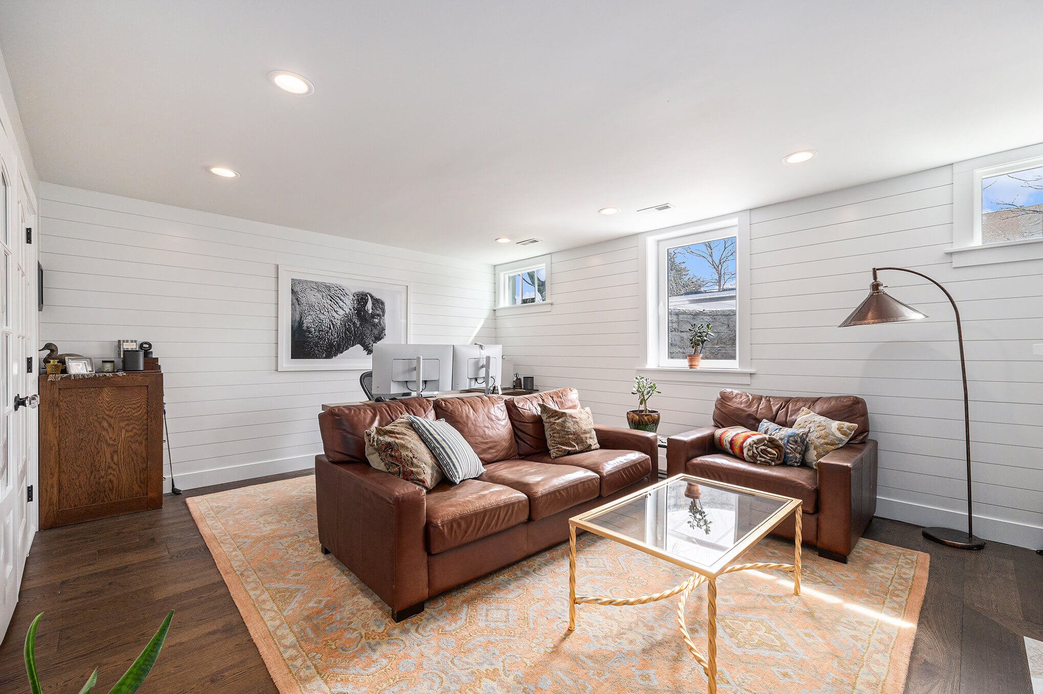 Cozy living room with brown leather sofas, a glass-topped coffee table, and a floor lamp. The room features white shiplap walls, large windows, and a decorative rug. A photo of a bison hangs on the wall.
