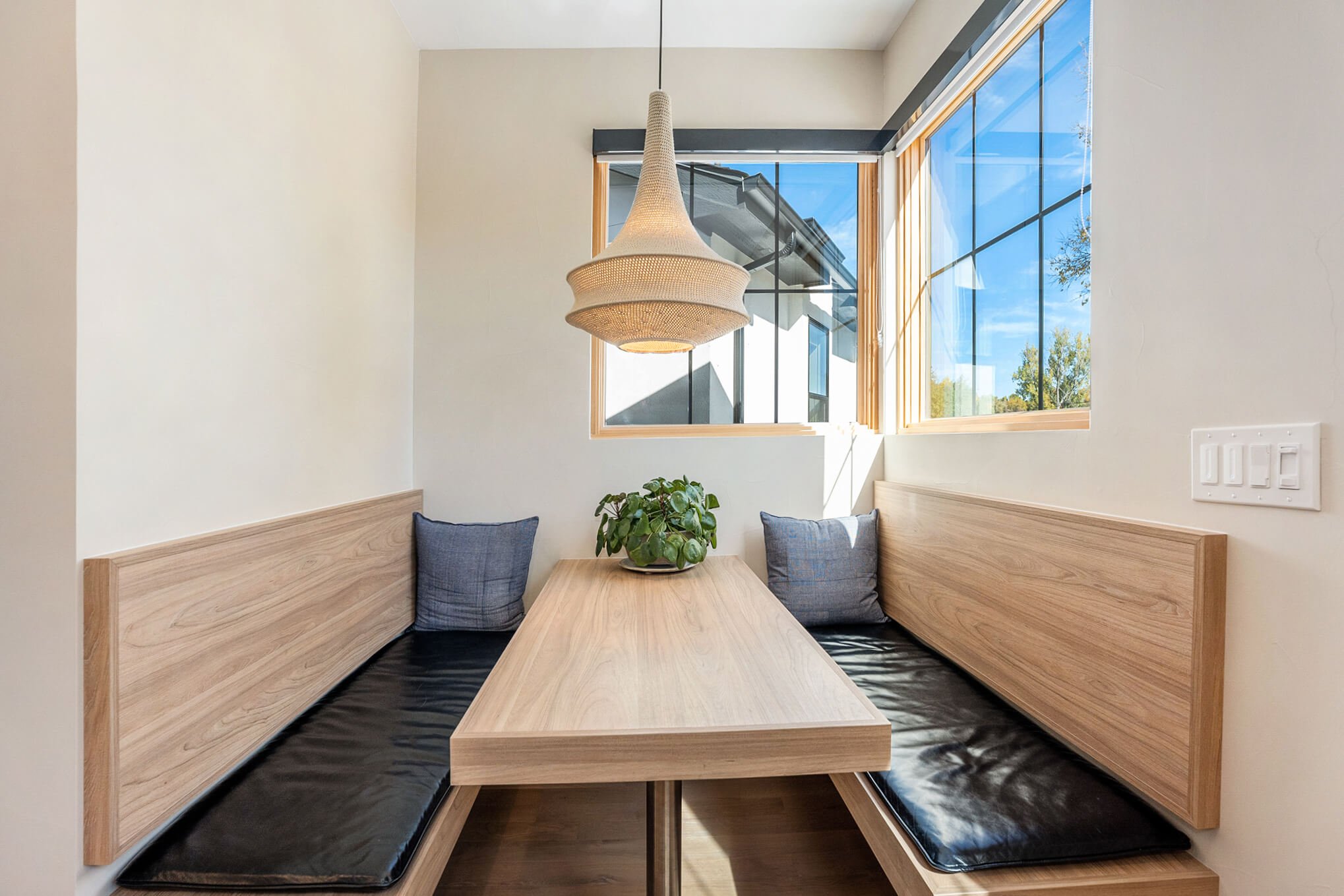 Modern dining nook with wooden benches and table, black cushions, gray pillows, a hanging light fixture, and a potted plant. Large windows provide natural light.