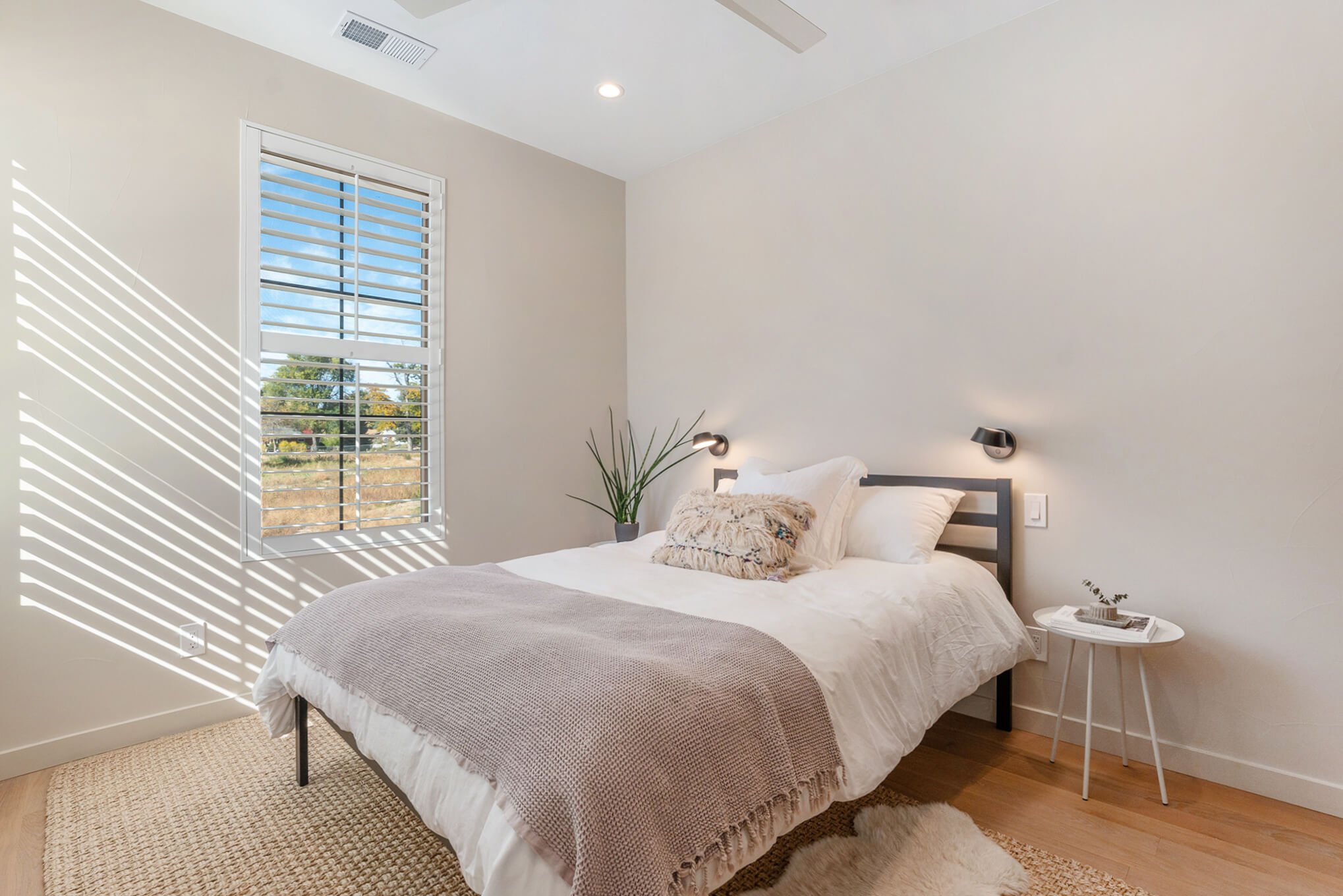Modern bedroom with a neatly made bed, beige throw blanket, decorative pillows, small side table, and window with blinds letting in natural light.