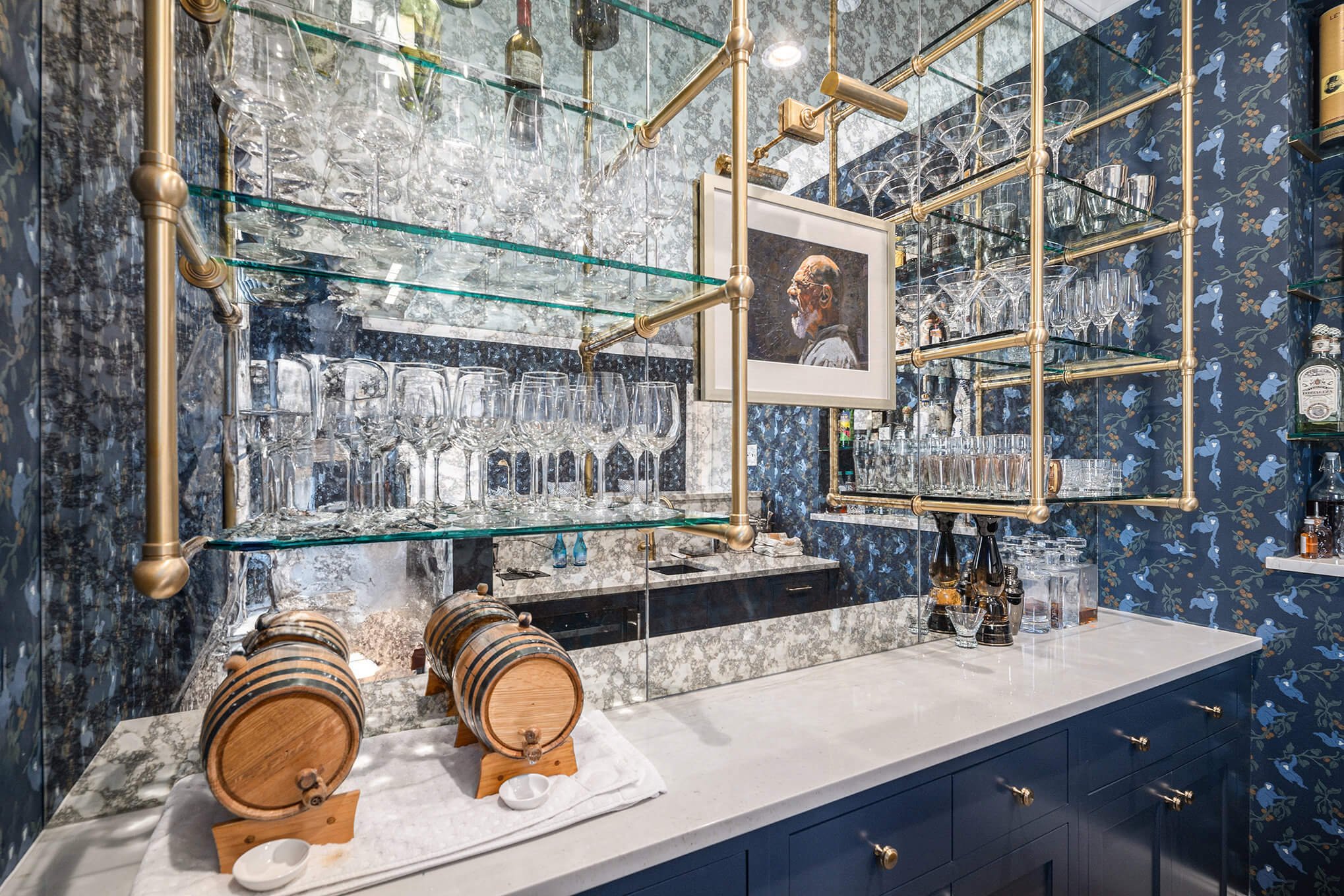 Home bar with glass shelving, wine glasses, liquor bottles, wooden barrels, and blue floral wallpaper.