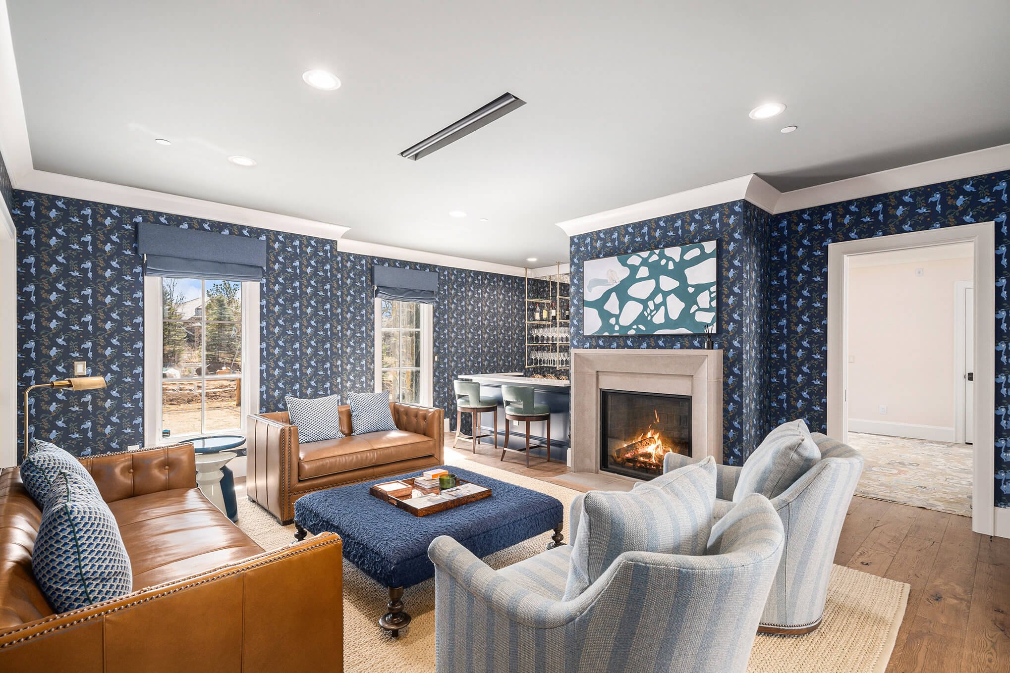 Cozy living room with blue patterned wallpaper, two brown leather sofas, a blue ottoman, light gray striped chairs, and a fireplace. Features a bar area with stools and decorative wall art.