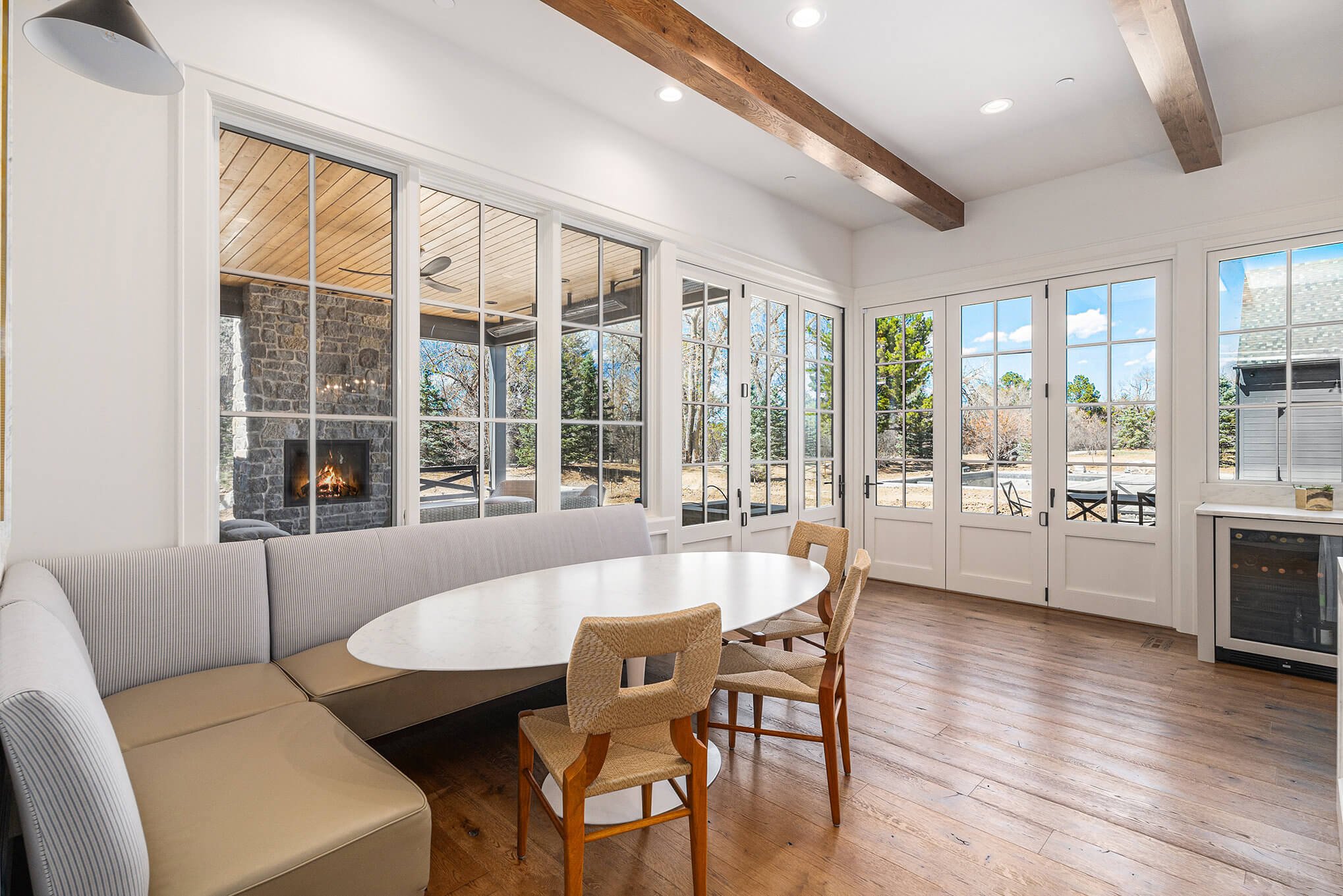 Modern dining area with oval table, beige cushioned bench, wooden chairs, and large windows overlooking outdoor patio with fireplace.