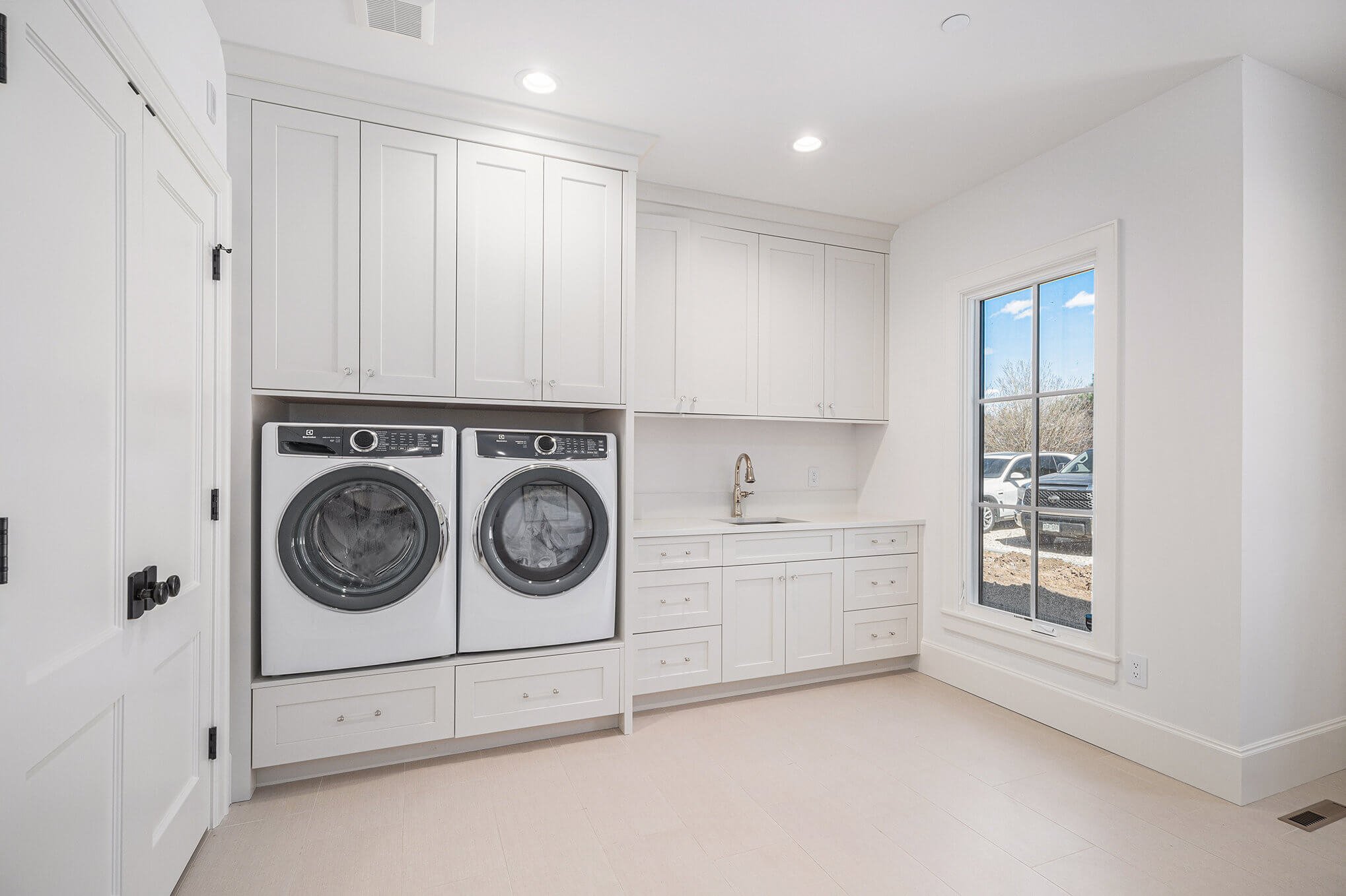 Modern laundry room with white cabinets, front-loading washer and dryer, sink, and large window.