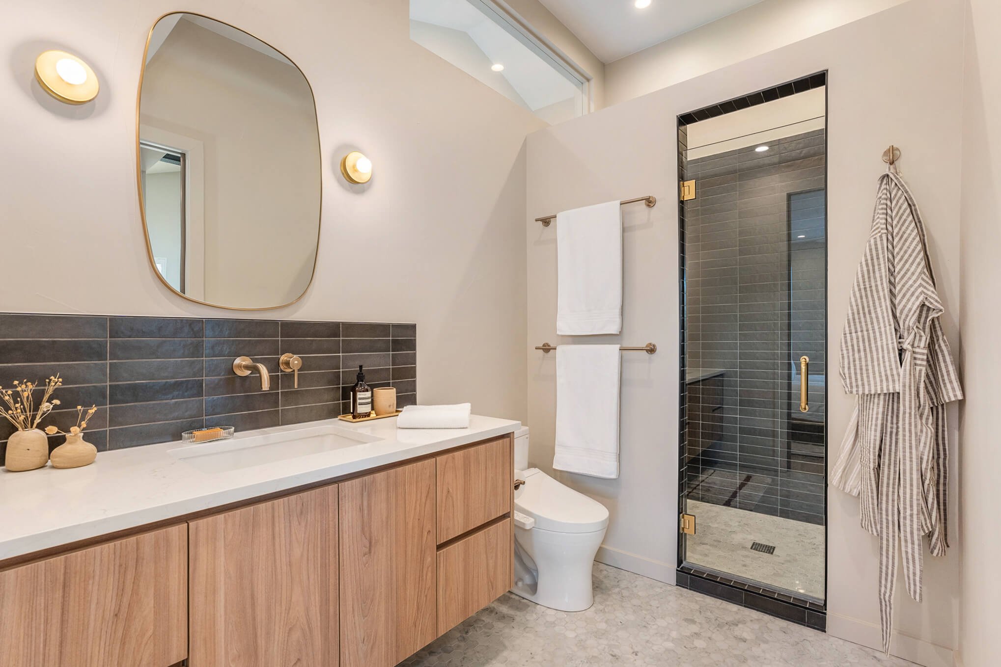 Modern bathroom with a wooden vanity, large mirror, two wall lights, dark subway tile backsplash, towel rack, toilet, and glass shower enclosure.
