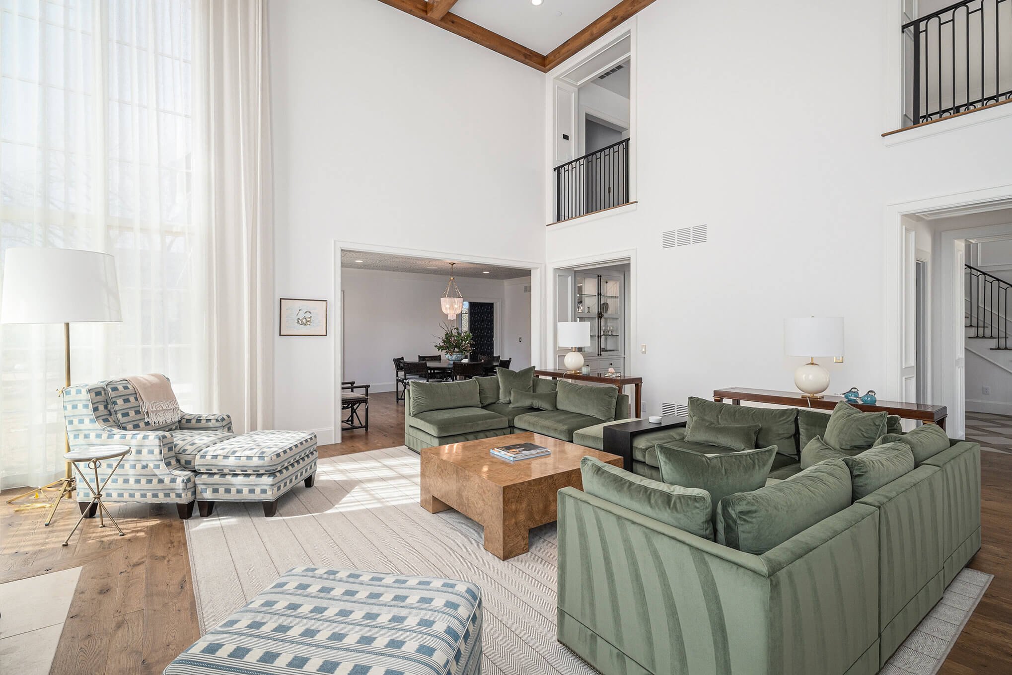 Spacious living room with high ceilings, featuring green sectional sofas, a wooden coffee table, a patterned armchair with an ottoman, and a large floor lamp. The room has wooden flooring, a tall window with sheer curtains, and an open doorway to a d