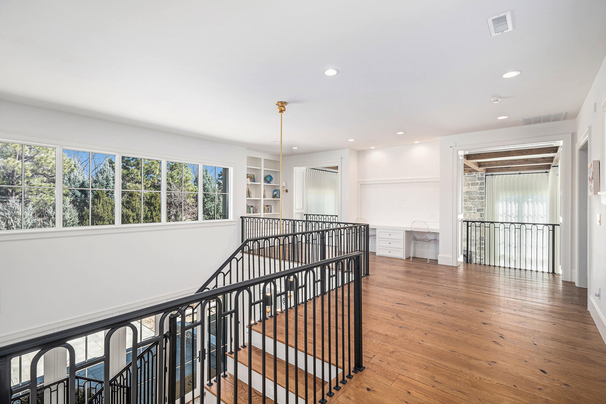Spacious second-floor landing with wooden flooring, black metal railings, large window displaying outdoor trees, built-in shelves with books and decor, desk with chair, and overhead lighting.