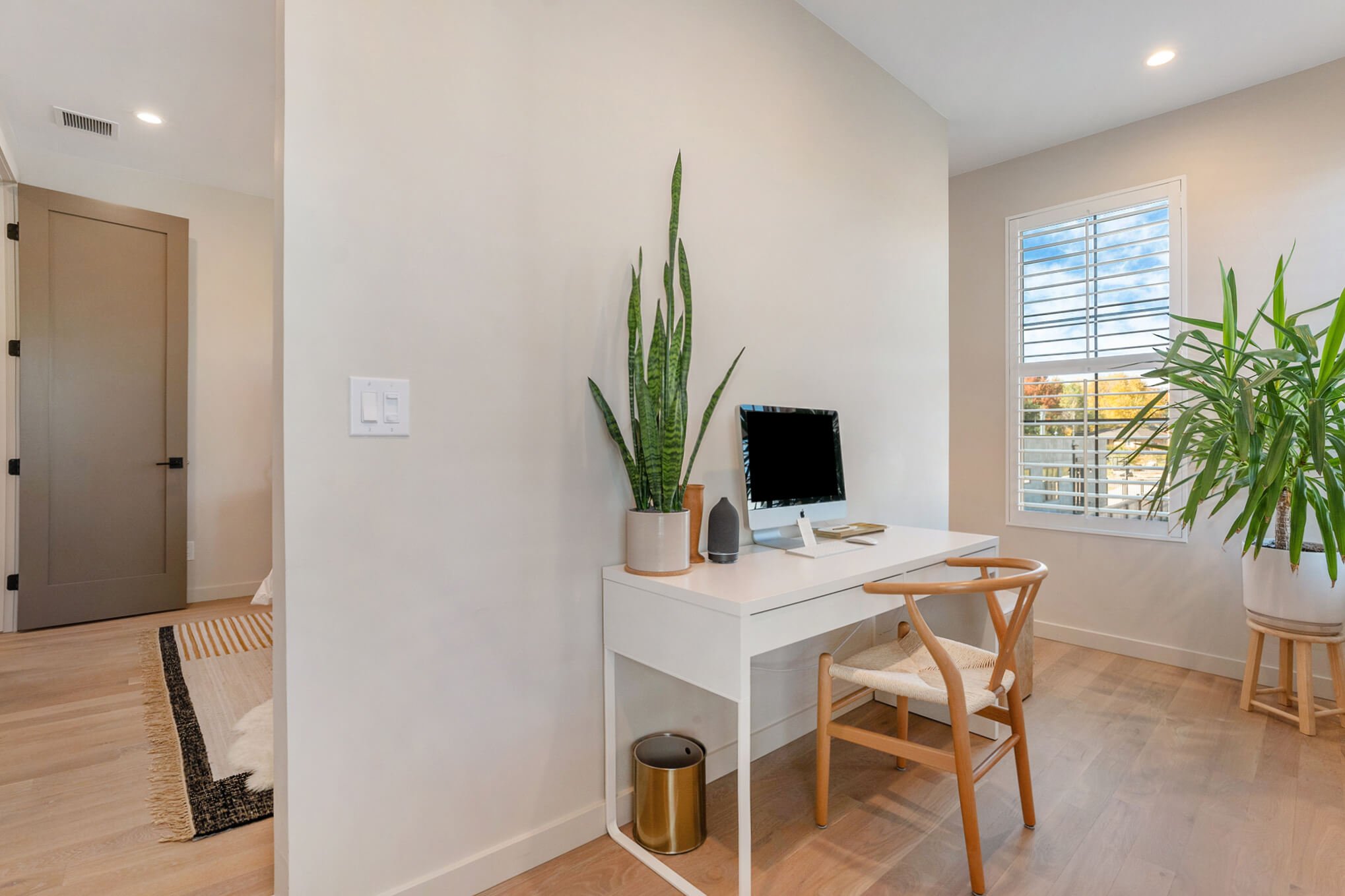 Modern home office with a white desk, computer, wooden chair, and plants, against a light-colored wall with a window.